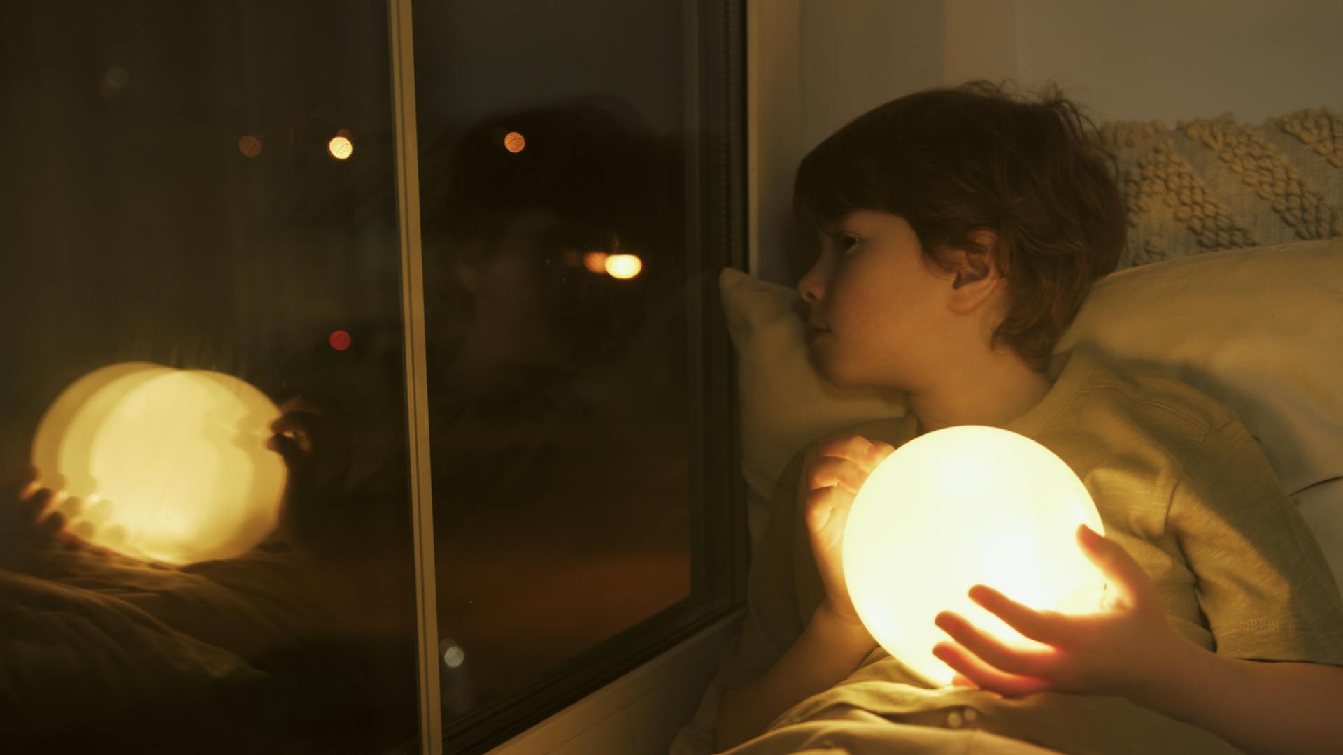Un jeune garçon tenant une boule de lumière qui regarde à travers sa fenêtre la nuit | Source : Pexels