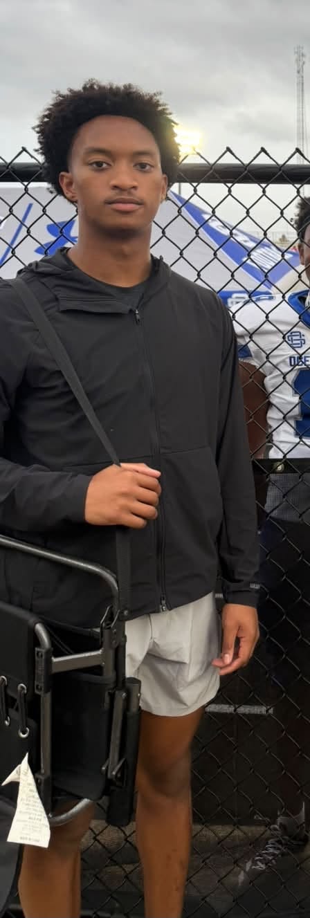 Vêtu d'une veste noire et d'un short, Nolan se tient près de la clôture pendant un match, son sac photo en bandoulière. | Source : Facebook/christine.wonsley