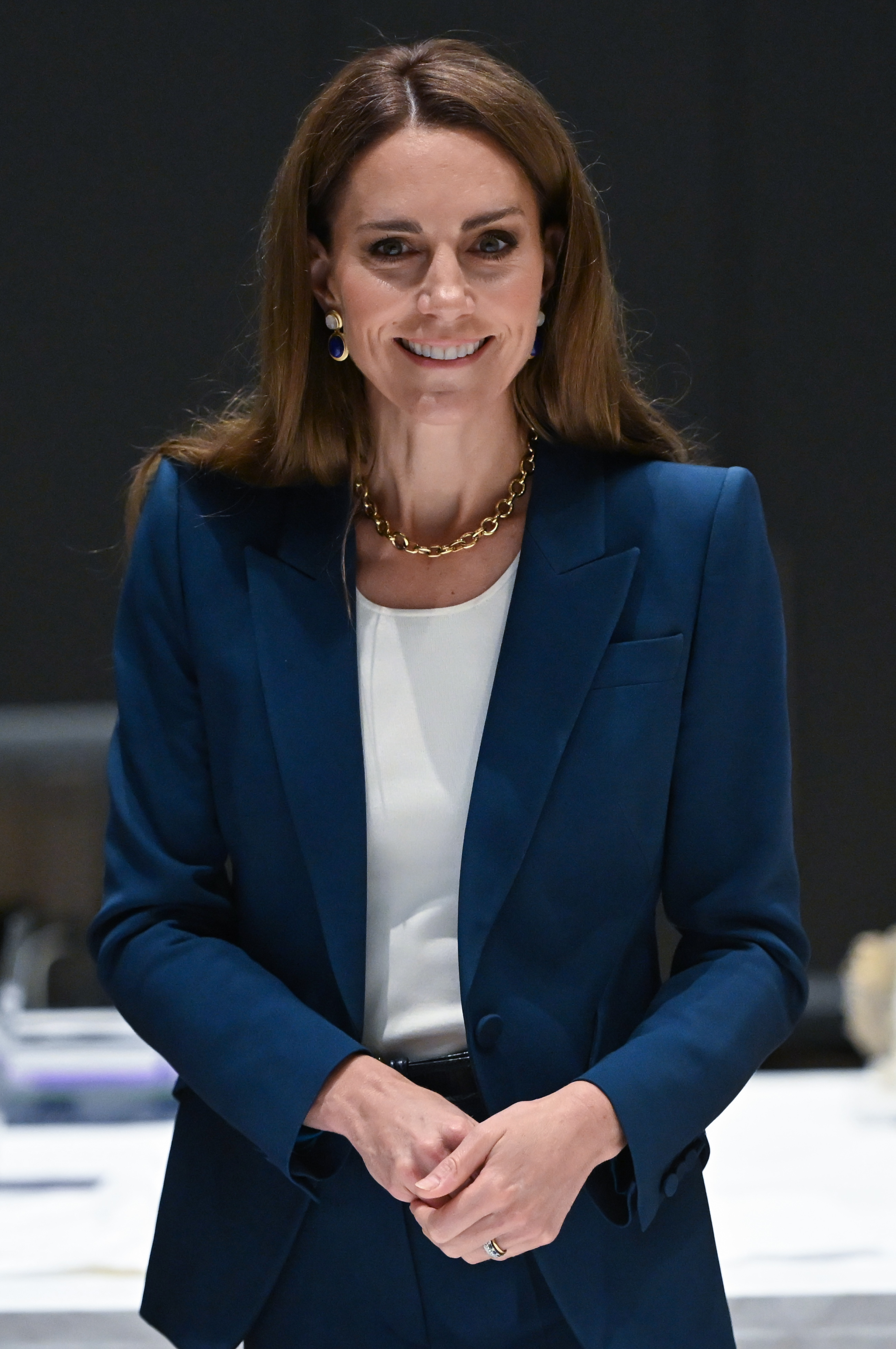 La princesse Catherine sourit doucement dans un tailleur bleu sur mesure et un haut blanc. Sa posture est droite, ses mains doucement jointes, elle rayonne d'une confiance calme alors qu'elle se tient à l'intérieur de l'entrepôt Est du V&A.