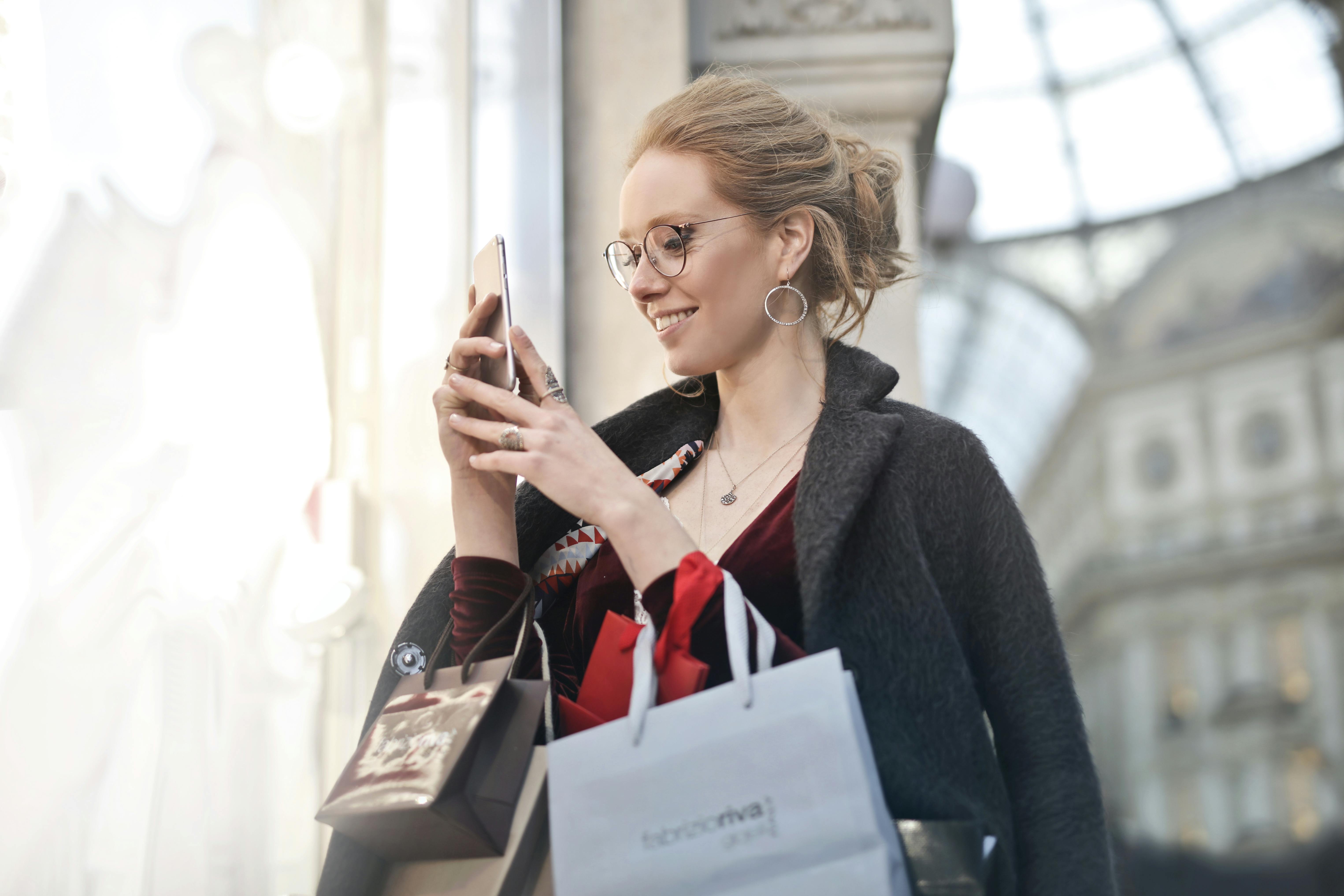 Une femme sourit en tenant plusieurs sacs à provisions tout en utilisant son téléphone | Source : Pexels