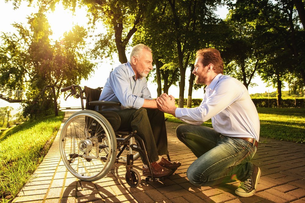 Un jeune homme et son vieux père en fauteuil roulant dans un parc. | Shutterstock