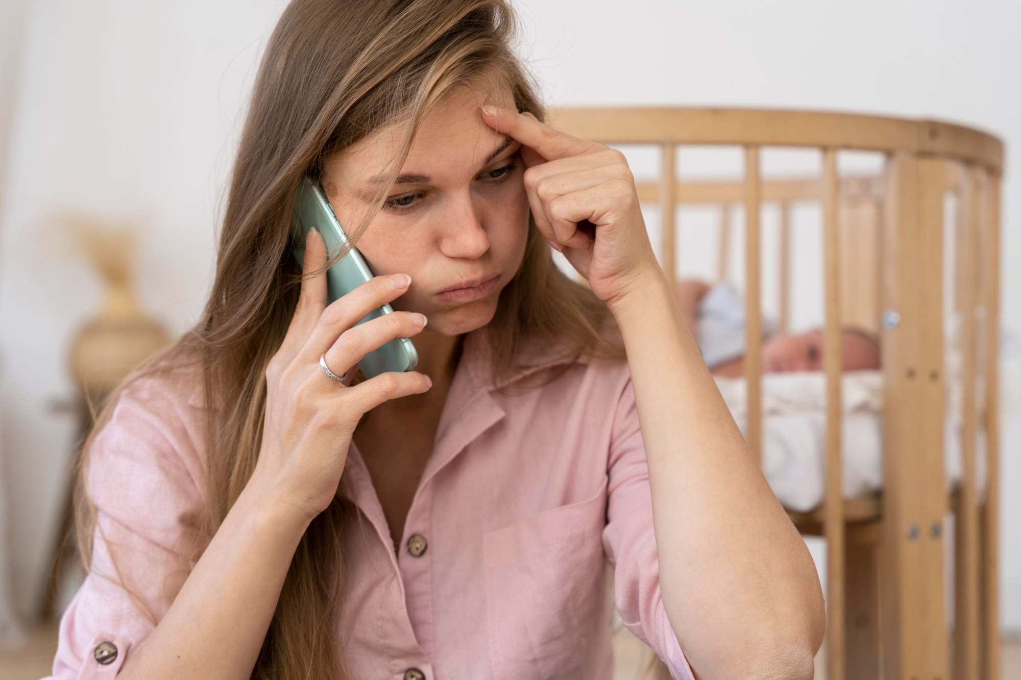 Une femme frustrée parle au téléphone avec son bébé couché dans un berceau derrière elle | Source : Freepik