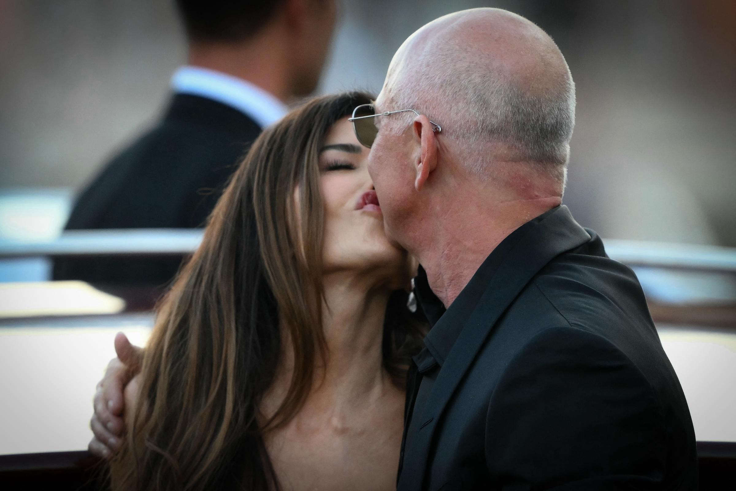 Lauren Sánchez et Jeff Bezos s'embrassent sous le regard d'un garde en costume, dans un moment de spontanéité qui se détache sur fond de bâtiments flous et de lumière du soir se reflétant sur l'eau.