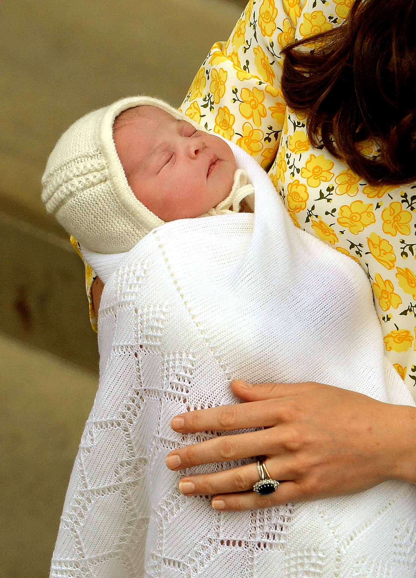 Catherine, duchesse de Cambridge, tient dans ses bras sa fille nouveau-née, la princesse Charlotte, devant l'aile Lindo de l'hôpital St Mary's, le 2 mai 2015, à Londres, en Angleterre. Le bébé endormi est emmailloté dans une douce couverture blanche en tricot, avec un bonnet assorti, et tenu bien au chaud par Catherine, qui porte une robe jaune à motifs floraux et sa bague de fiançailles en saphir. | Source : Getty Images