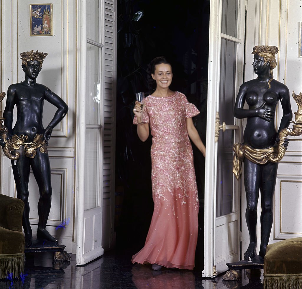 Jeanne MOREAU, la muse de Pierre Cardin, en robe longue brodée de brillants, tenant un verre, dans l'entrée d'un salon encadrée de deux statues porte-flambeau. | Photo : Getty Images