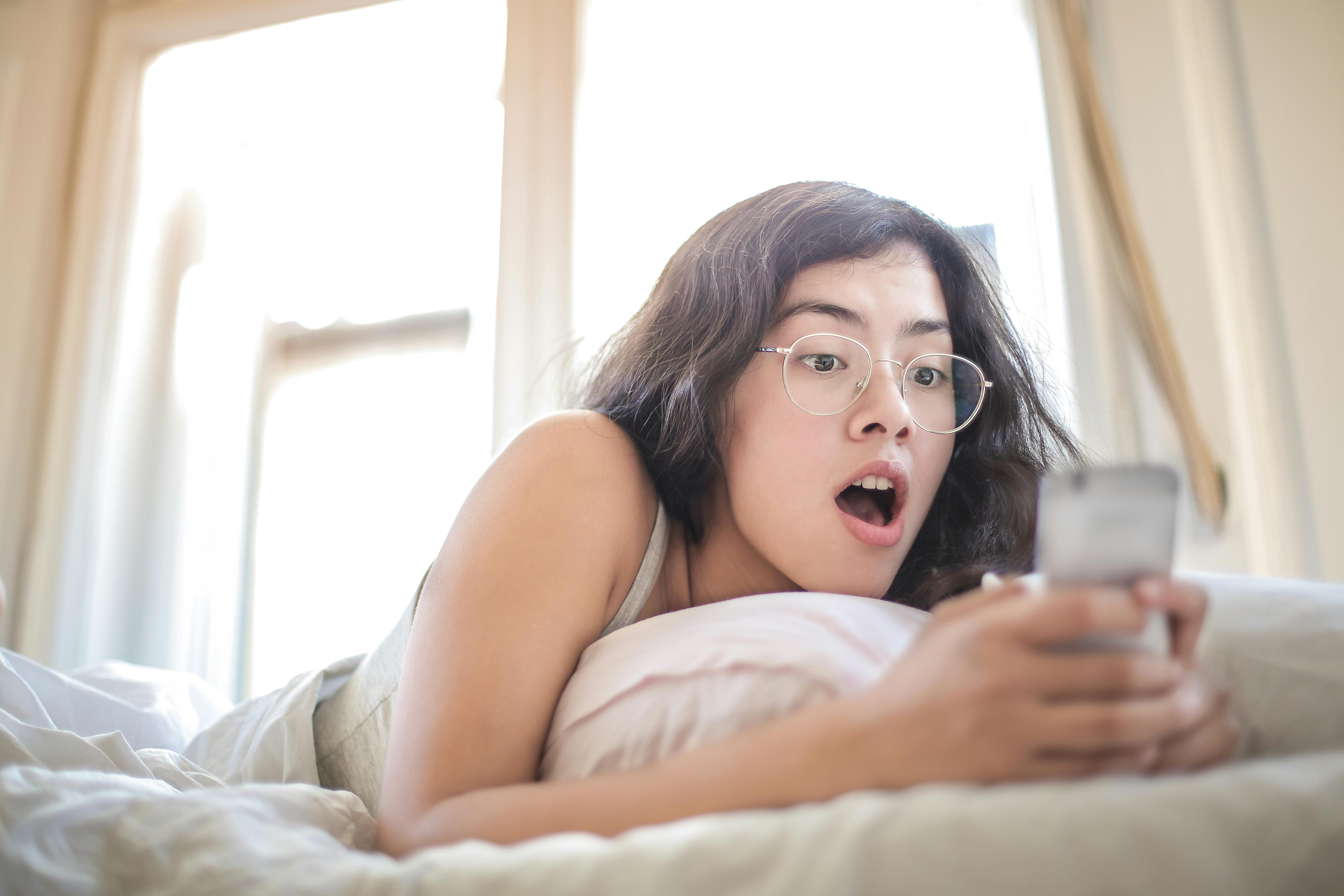 Une femme choquée, allongée sur un lit, tenant et regardant un téléphone | Source : Pexels