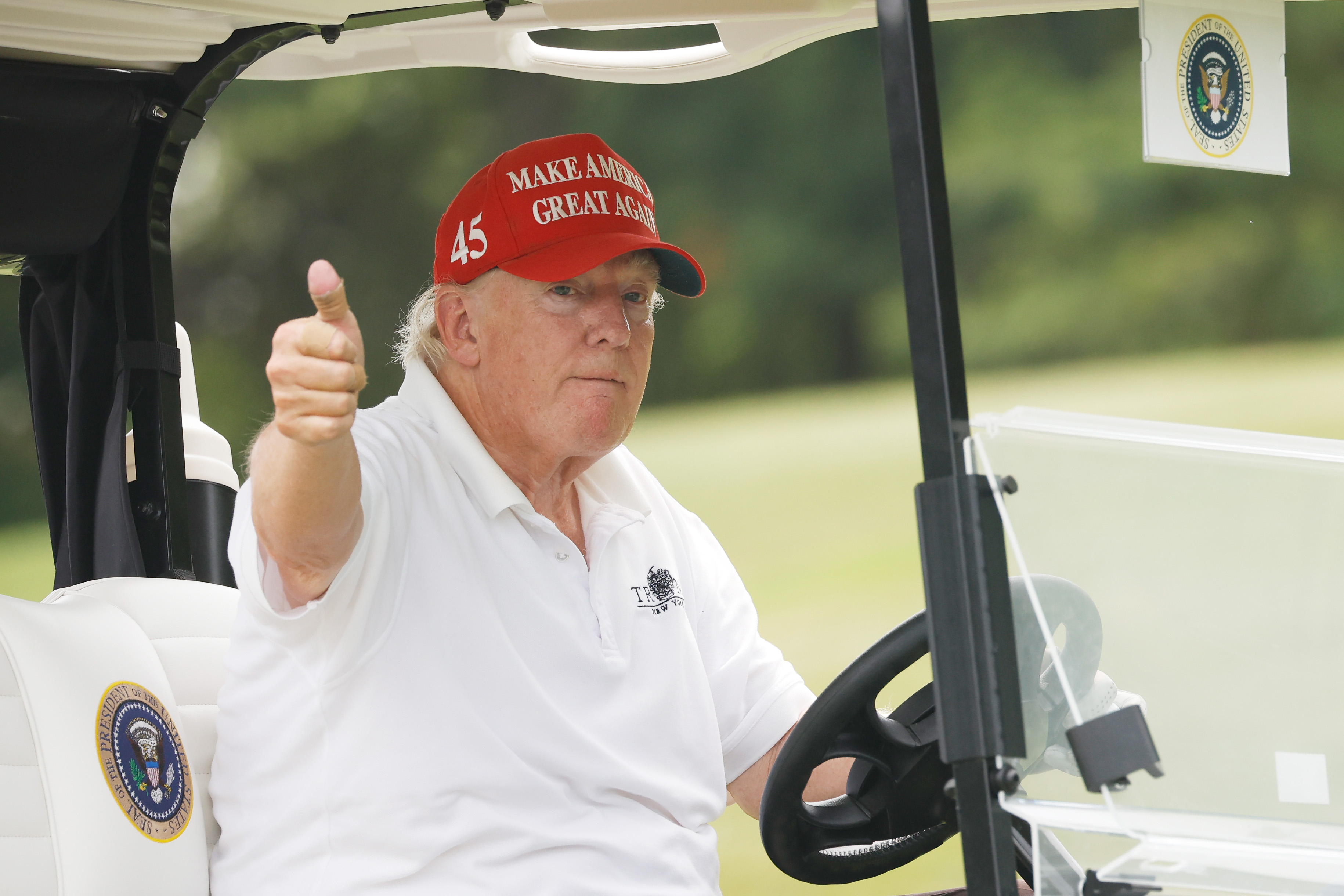 Assis dans une voiturette de golf, Donald Trump lève le pouce d'un air sérieux. La différence de couleur entre son visage et son cou est visible sous la lumière du jour.