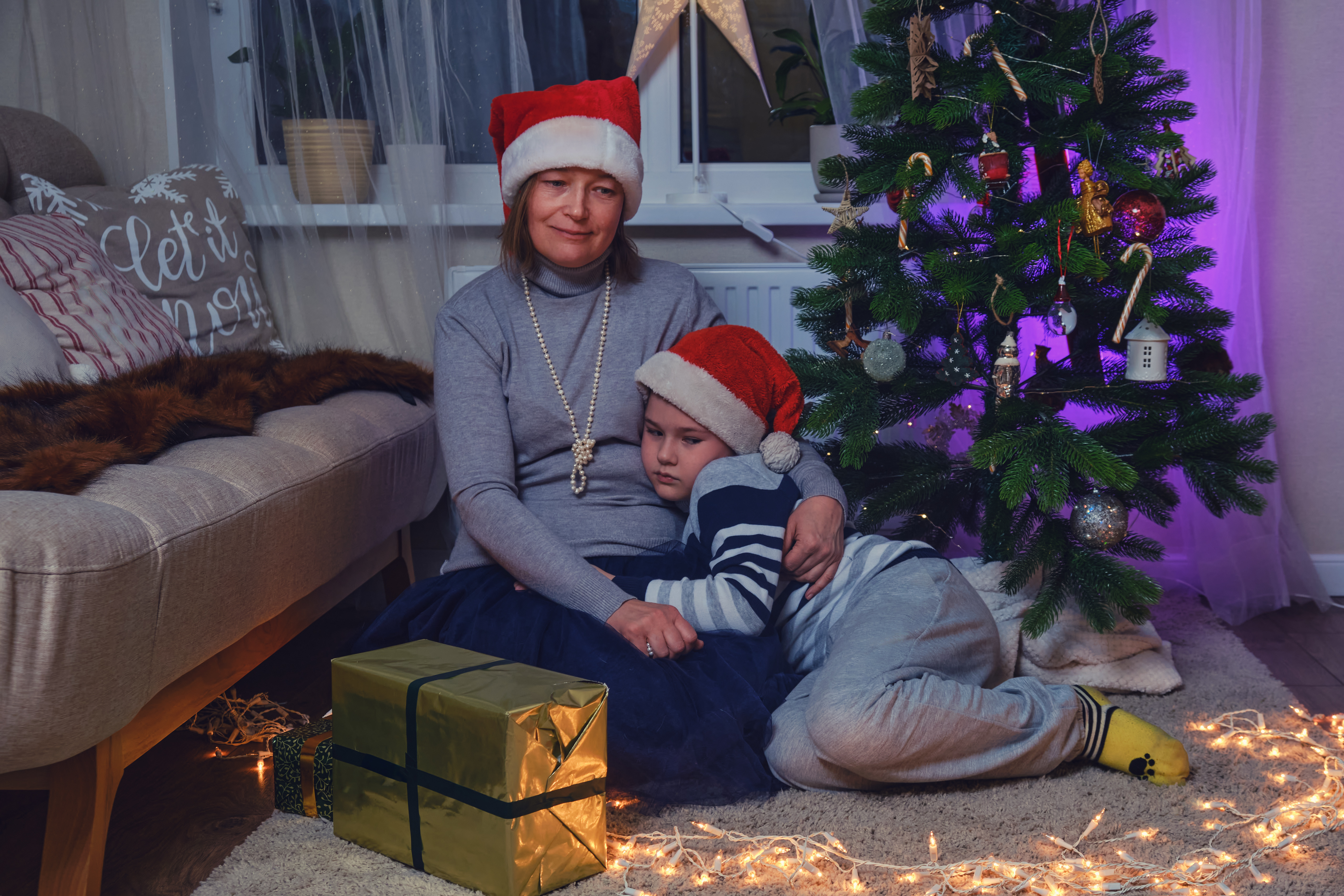 Mère et fils tristes assis près du sapin de Noël dans les guirlandes lumineuses de la nuit. | Source : Getty Images