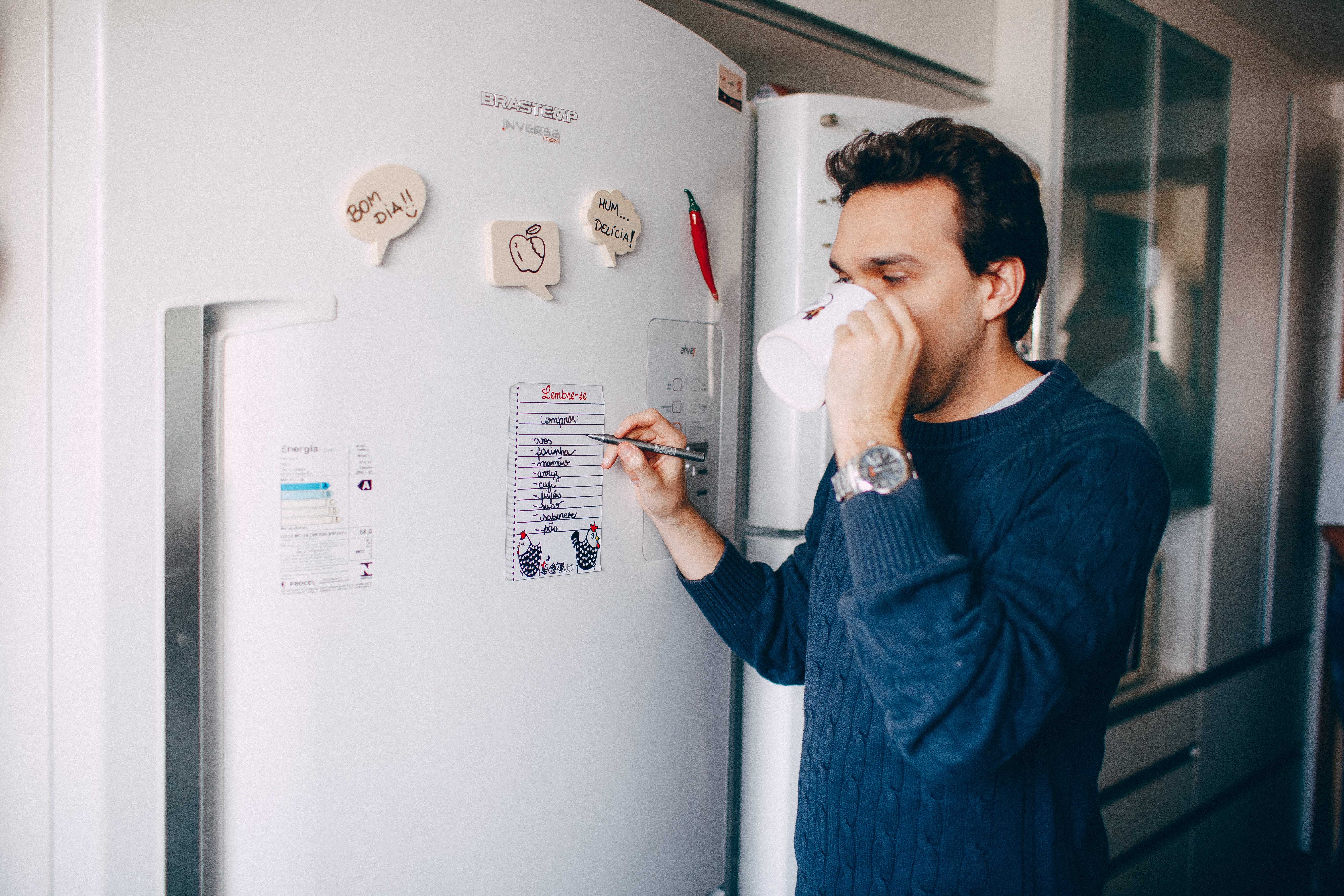 Un homme boit dans sa tasse tout en écrivant quelque chose sur un papier collé sur un réfrigérateur | Source : Pexels