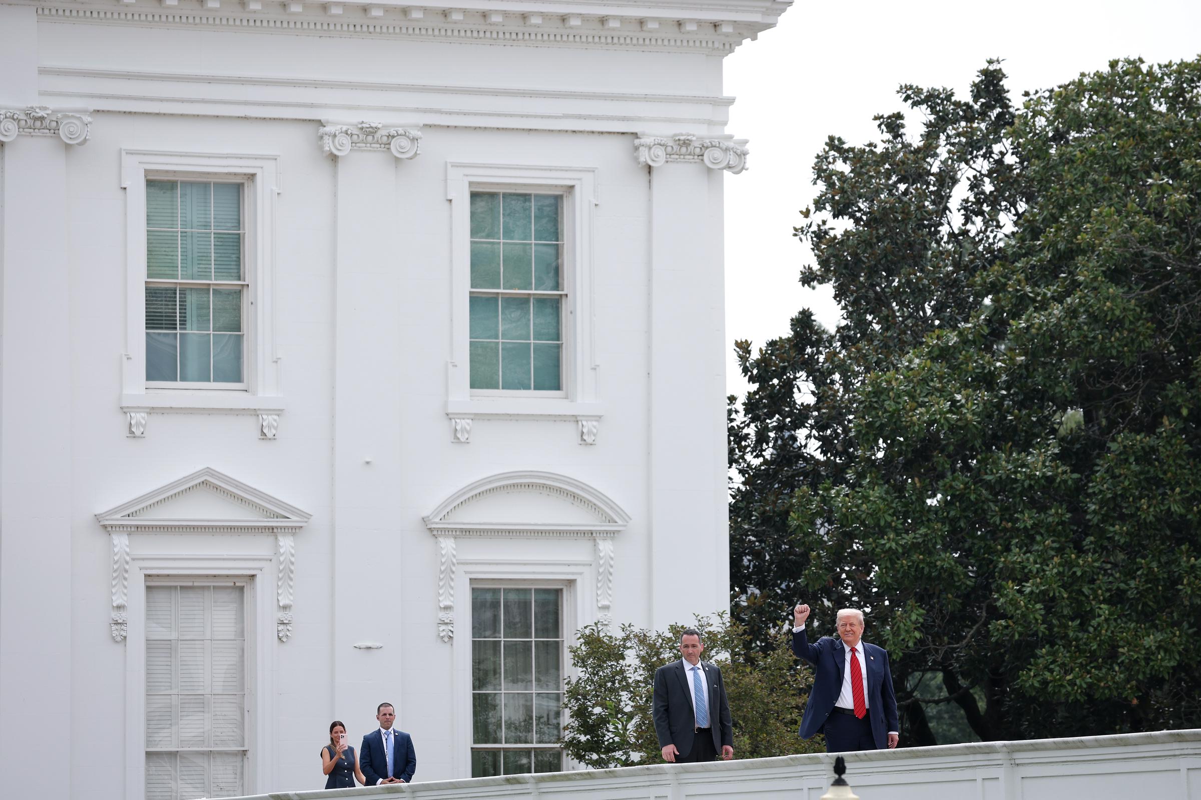 Donald Trump semble faire un signe de la main alors qu'il s'adresse aux journalistes lors d'une visite sur le toit de l'aile ouest de la Maison Blanche à Washington, rejoint par des assistants et du personnel de sécurité.