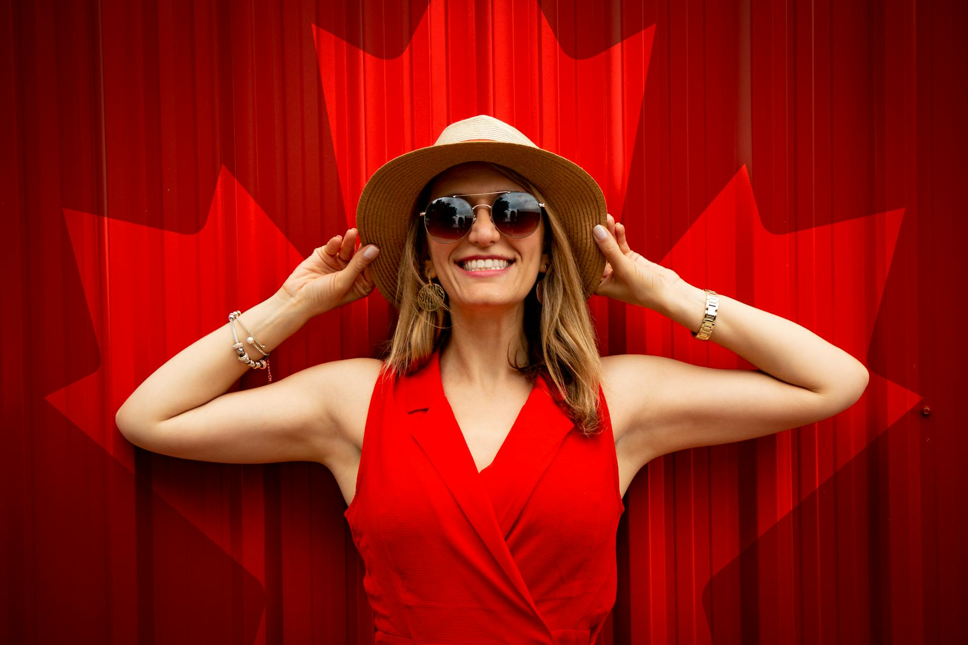 Une femme souriante vêtue d'une robe rouge, de lunettes de soleil et d'un chapeau, debout devant une toile de fond rouge | Source : Pexels