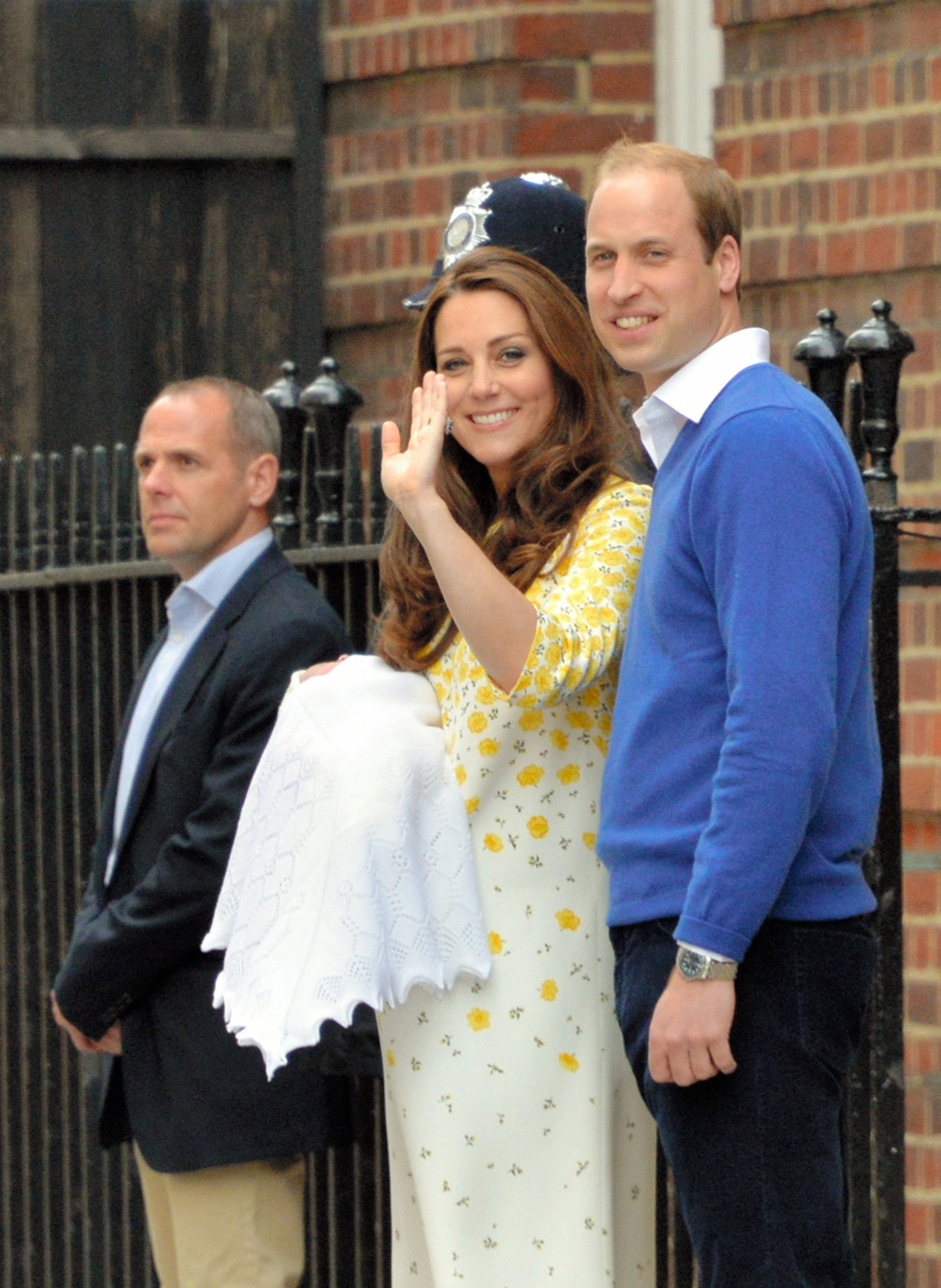Le prince William, duc de Cambridge, sourit fièrement aux côtés de Catherine, duchesse de Cambridge, tandis qu’elle salue la foule en liesse rassemblée devant les marches de l’établissement médical. William porte un pull bleu roi vif à col en V par-dessus une chemise blanche à col ouvert et un pantalon bleu marine foncé. Il se tient près des agents de sécurité tandis que le couple présente au public le nouveau membre de la famille royale. | Source : Getty Images