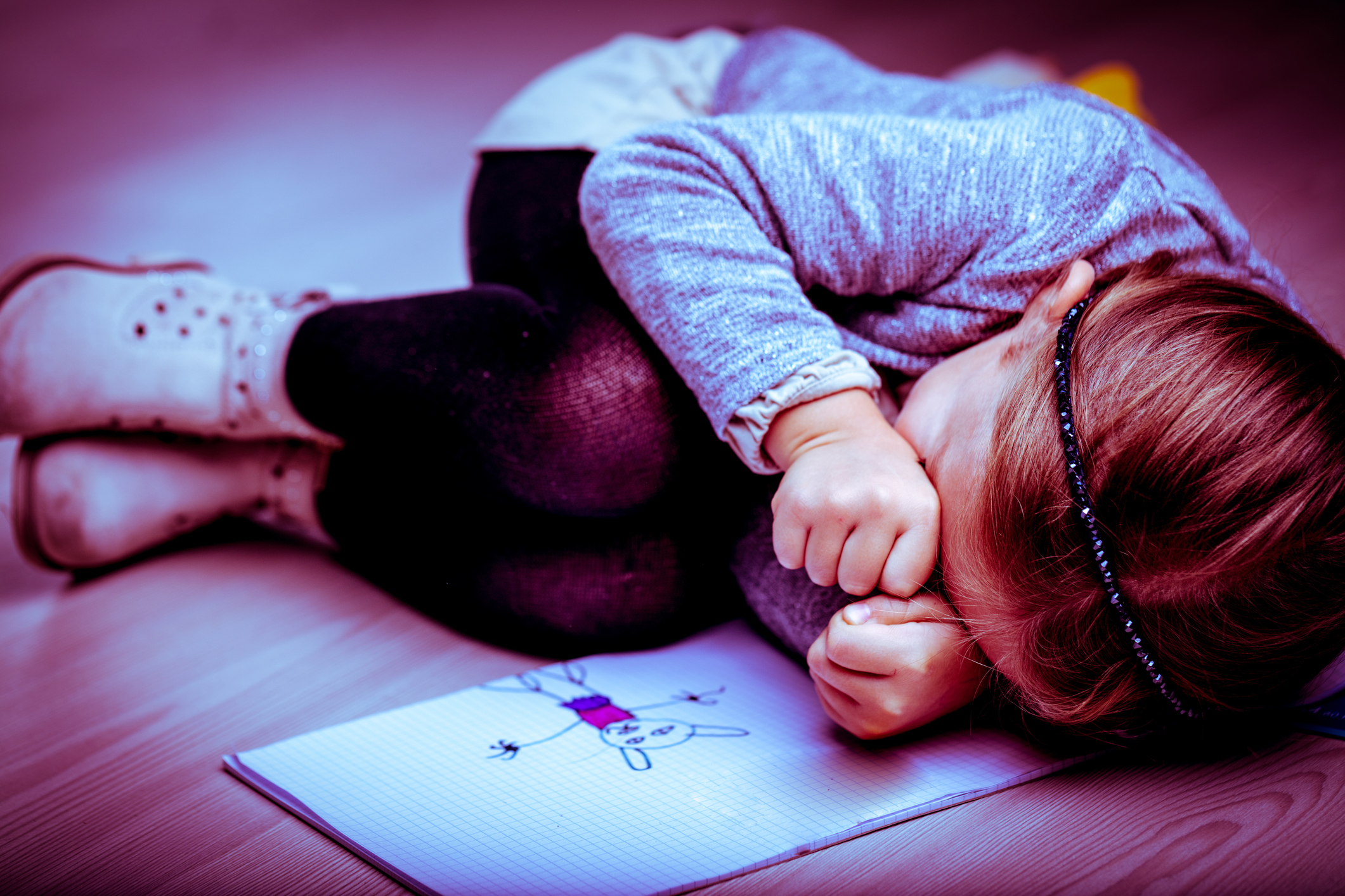 Petite fille bouleversée, recroquevillée sur le lit à côté de son dessin, se frottant les yeux avec les poings comme si elle pleurait I Photo d'illustration I Source : Getty Images