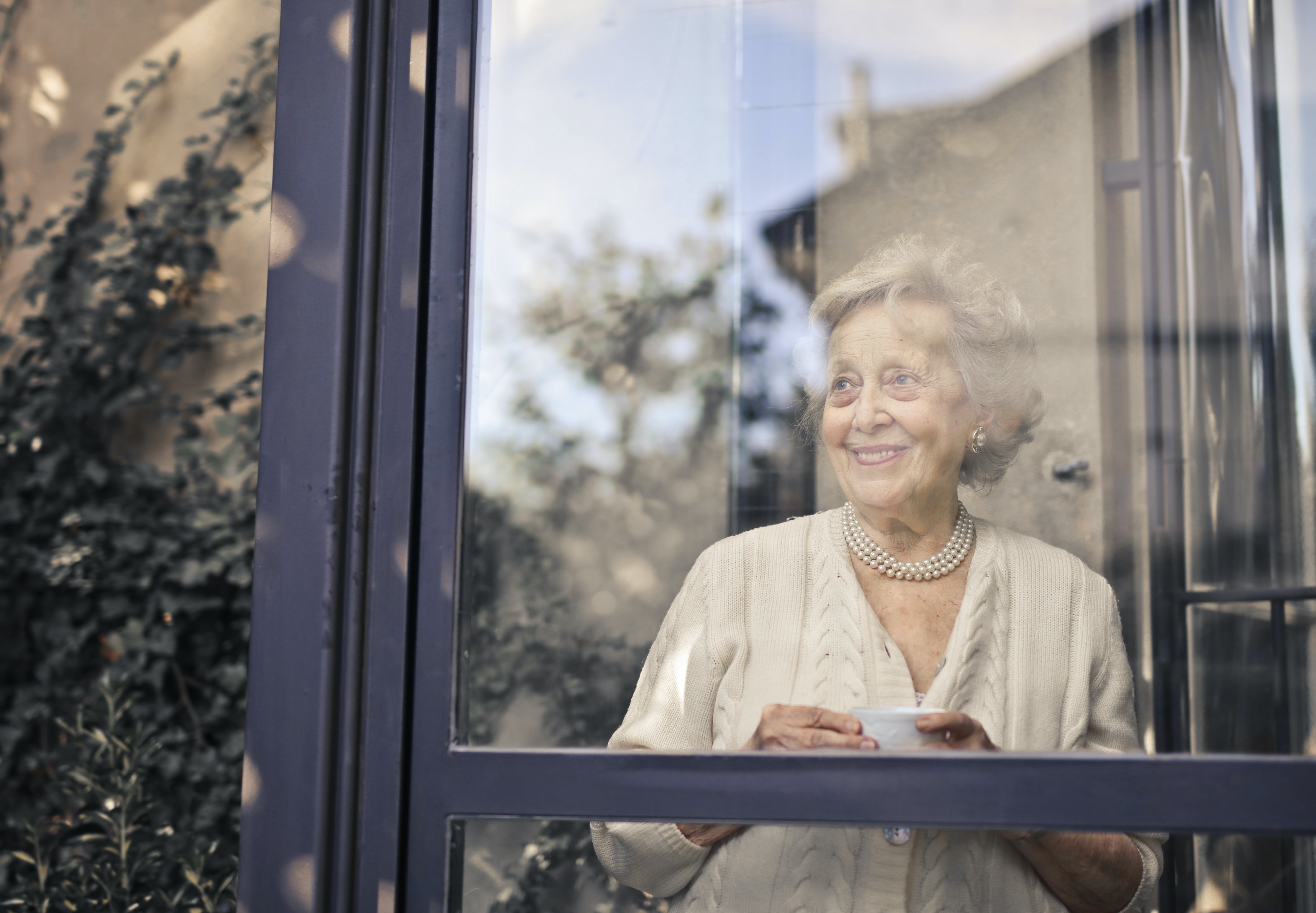 Une femme âgée sourit en tenant une boisson et en regardant par la fenêtre | Source : Pexels