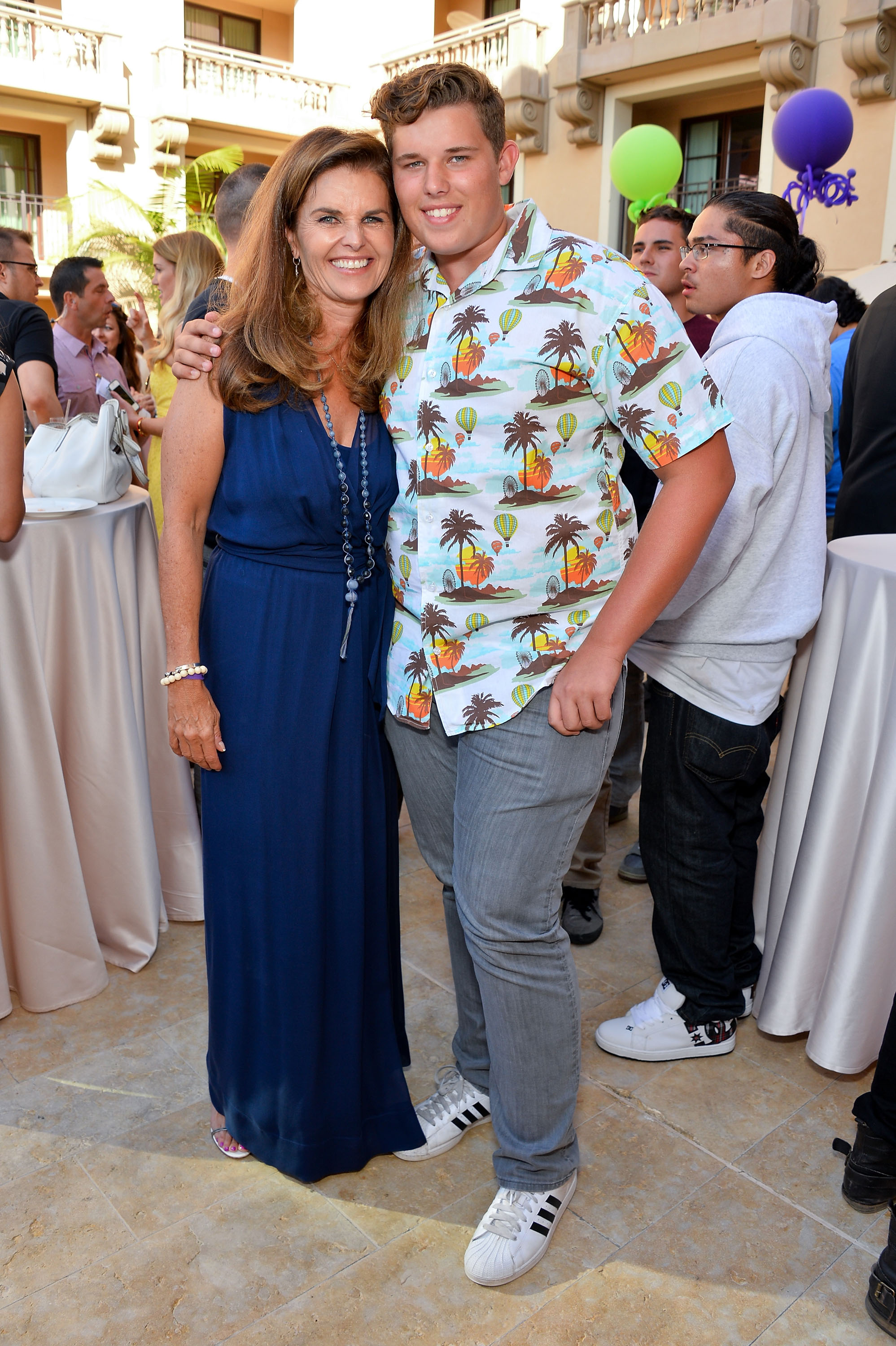 Maria Shriver et son fils Christopher partagent un moment chaleureux lors du même événement. Shriver porte une robe bleu profond avec des colliers superposés, tandis que Christopher arbore sa chemise imprimée caractéristique et un jean gris. Ils se tiennent près l'un de l'autre, entourés d'invités dans une ambiance festive.