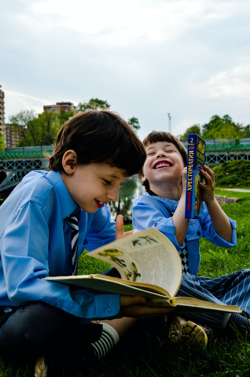 Deux garçons assis sur l'herbe en riant et en lisant des livres | Source : Pexels