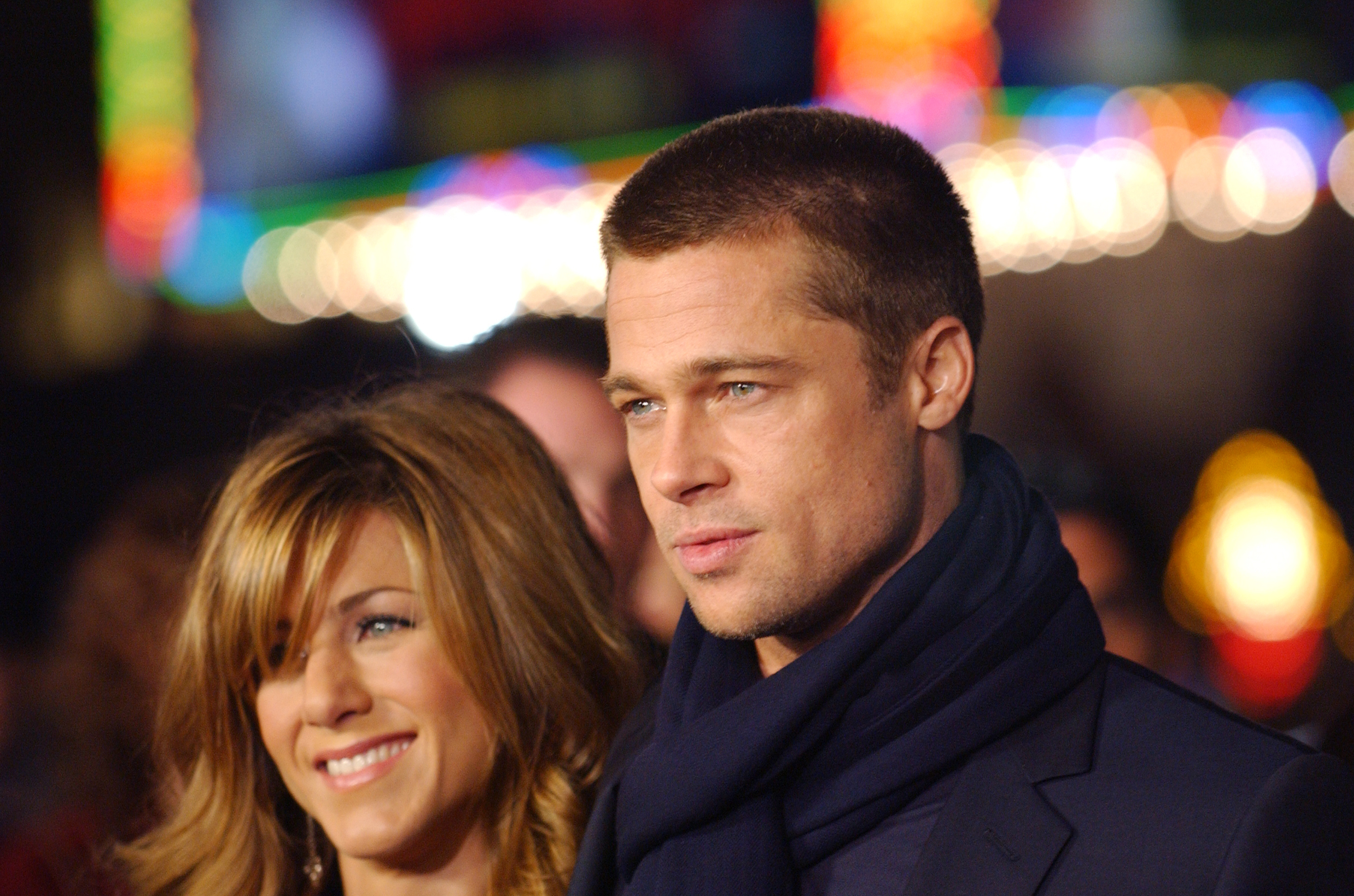 Jennifer Aniston sourit aux côtés de Brad Pitt lors de la première du film « Along Came Polly » à Los Angeles, avec en arrière-plan des lumières colorées et une foule floue qui scintille derrière le couple.