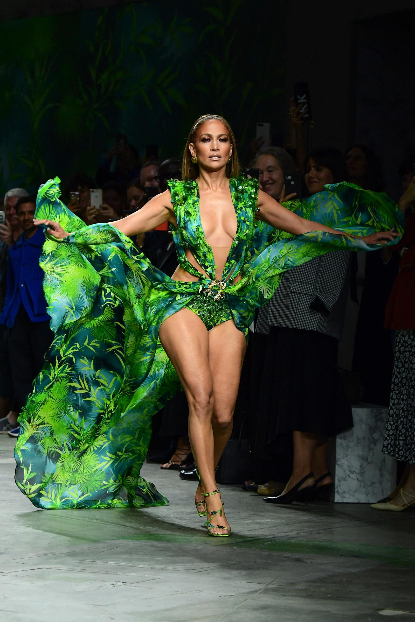 Jennifer Lopez domine la piste dans un look Versace dramatique à l'imprimé tropical, tendant les deux bras vers l'extérieur alors qu'un tissu vert fluide se déploie derrière elle. La tenue plongeante, les talons assortis et la foulée puissante transforment l'apparition au défilé de mode en un moment théâtral.