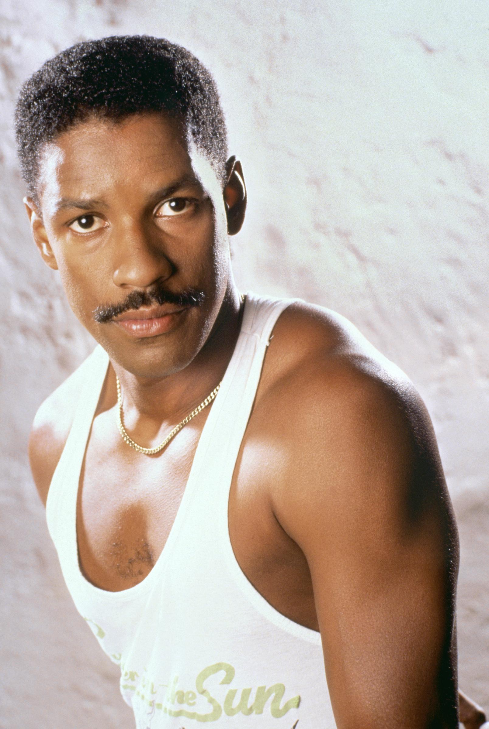 Denzel Washington respire la confiance dans ce portrait où il pose vêtu d'un débardeur blanc et d'une chaîne en or. Avec ses cheveux courts et sa moustache, son look est soigné et élégant, un style qui est devenu l'un de ses looks les plus reconnaissables à ses débuts.