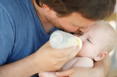 Un adorable bébé buvant du lait par le biais d'un biberon entre les mains de son père. | Shutterstock