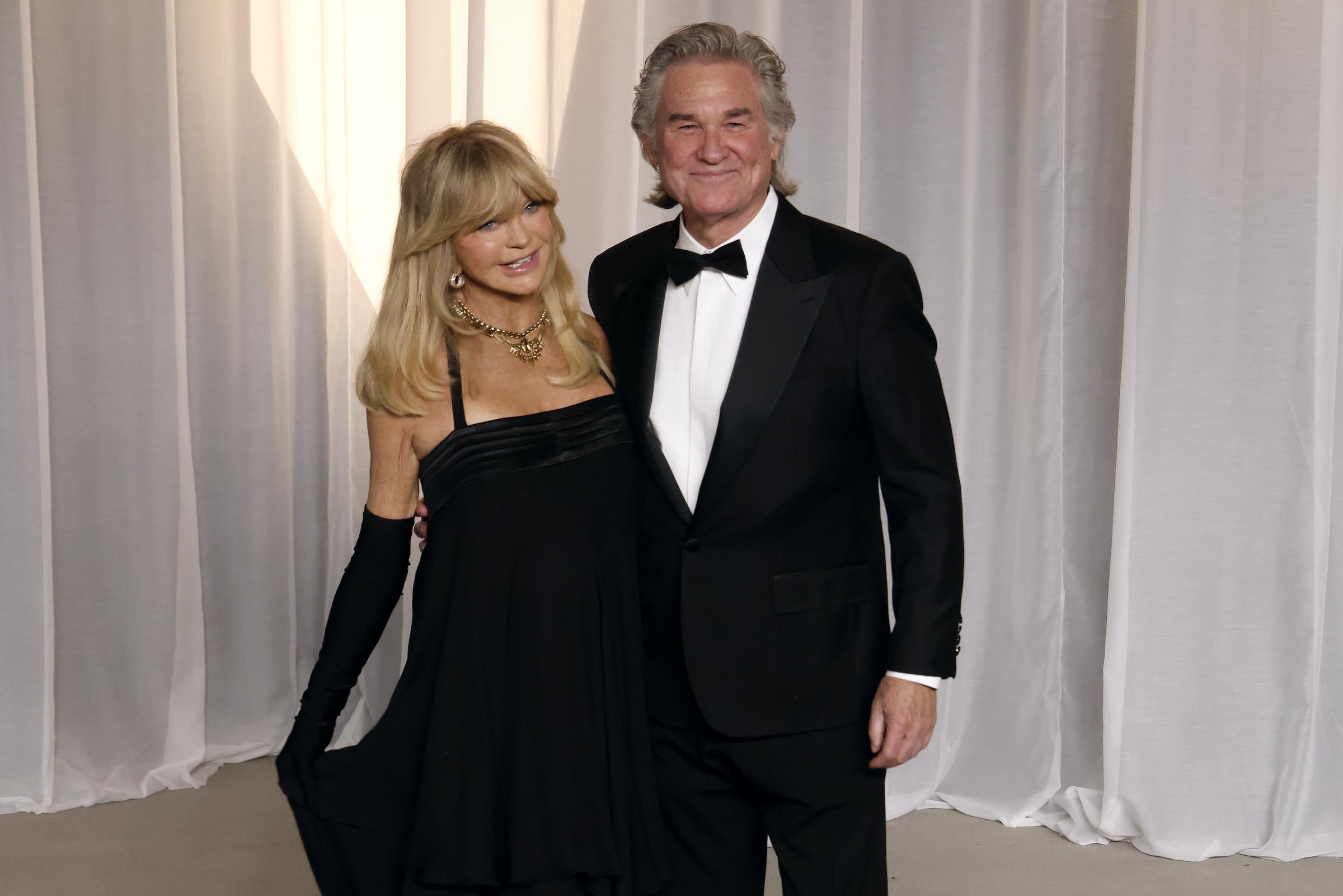 Goldie Hawn et Kurt Russell forment un couple naturellement chic dans leurs tenues de soirée noires assorties. Hawn est ravissante dans une robe de cocktail noire à taille empire, qu’elle porte avec des gants longs assortis, des bijoux en or qui en jettent et sa célèbre frange blonde volumineuse. Russell complète son style classique avec un smoking noir impeccable, doté de revers en satin en pointe, d’une chemise blanche impeccable et d’un nœud papillon noir traditionnel.