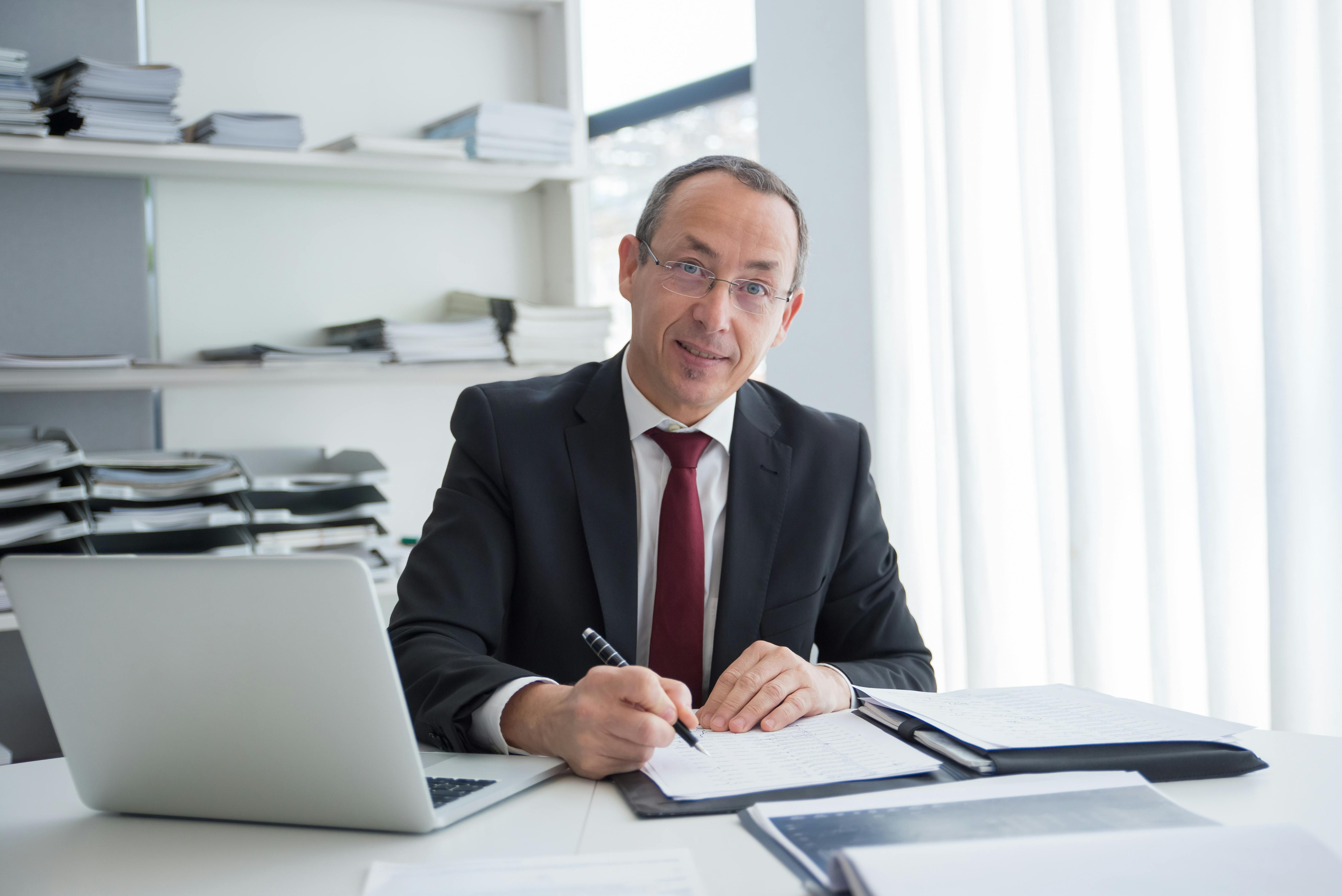 Un homme souriant assis avec un ordinateur portable et quelques documents | Source : Pexels