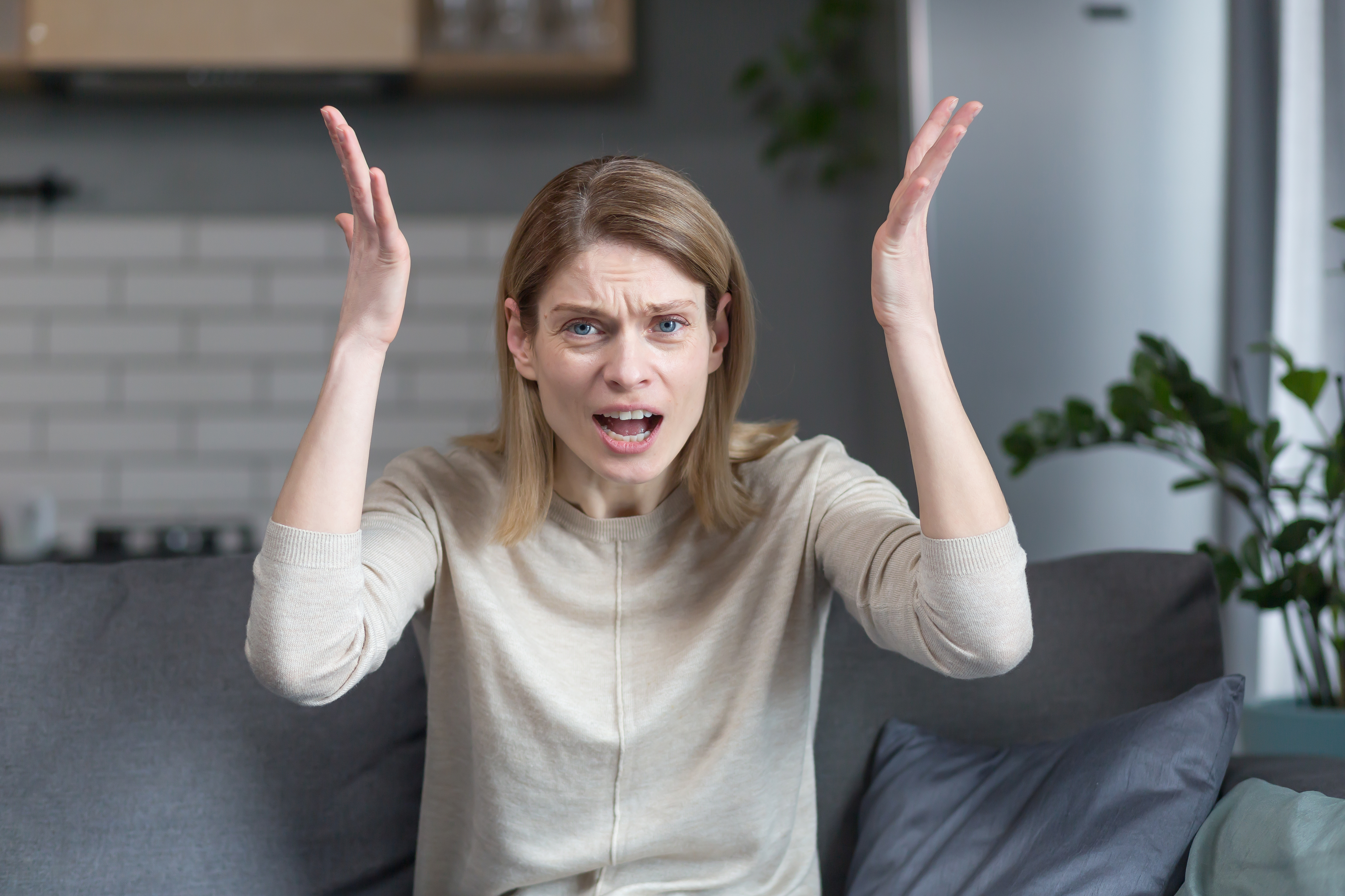 Une femme en colère et frustrée parle lors d'un appel vidéo en regardant la webcam, faisant des gestes furieux avec les mains alors qu'elle est assise sur le canapé à la maison | Source : Getty Images