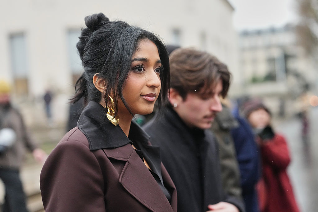 Maitreyi Ramakrishnan porte ses cheveux noirs en un long chignon relevé avec une raie au milieu, de grandes boucles d'oreilles en or, une chemise noire à large col sous un blazer en laine marron à larges revers, lors de la Fashion Week de Paris - Mode masculine automne/hiver 2026-2027, le 25 janvier 2026 à Paris, France I Source : Getty Images