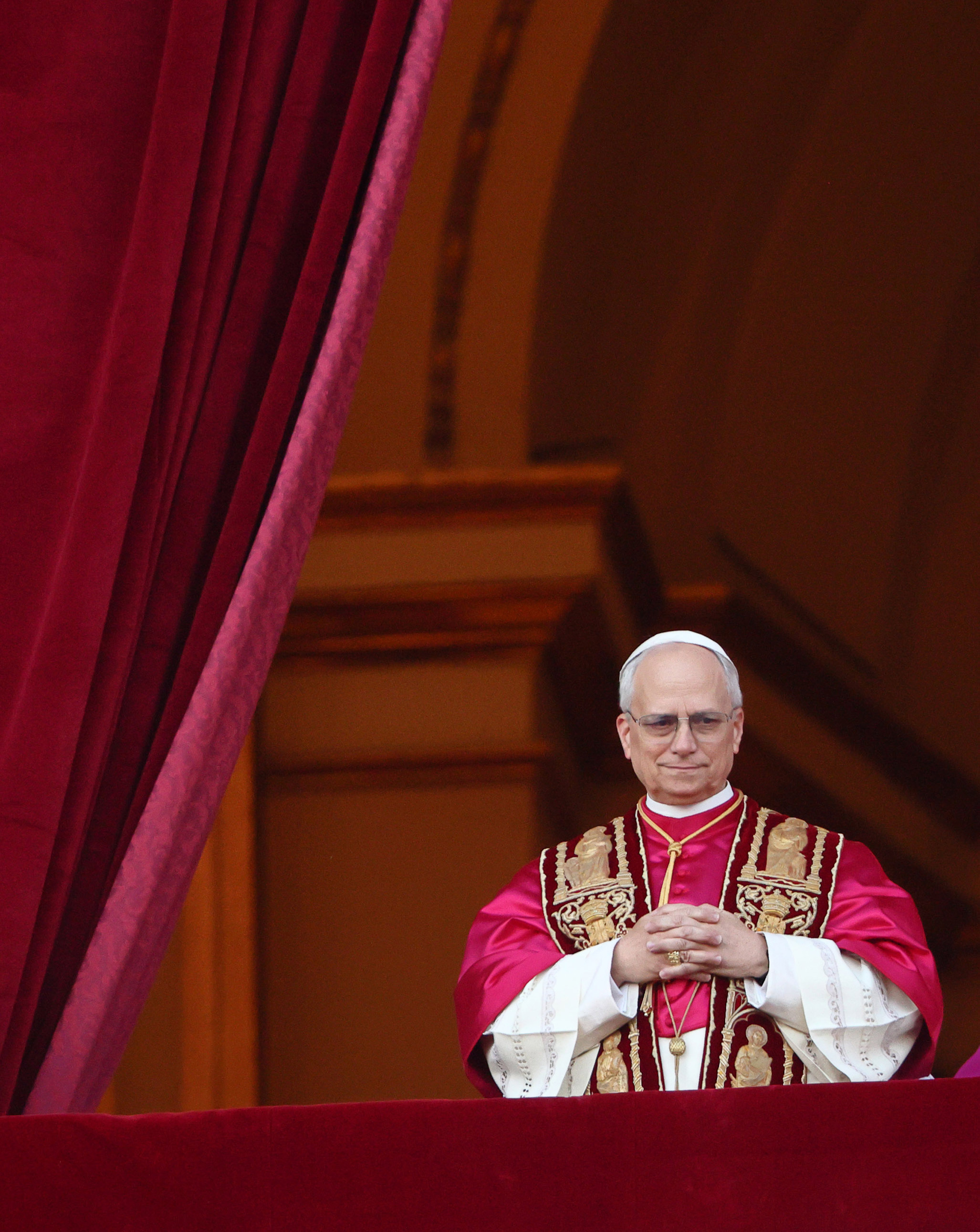 Le pape Léon XIV se tient solennellement entre de riches draperies de velours, les mains jointes au-dessus d'ornements cramoisis brodés de saints dorés.