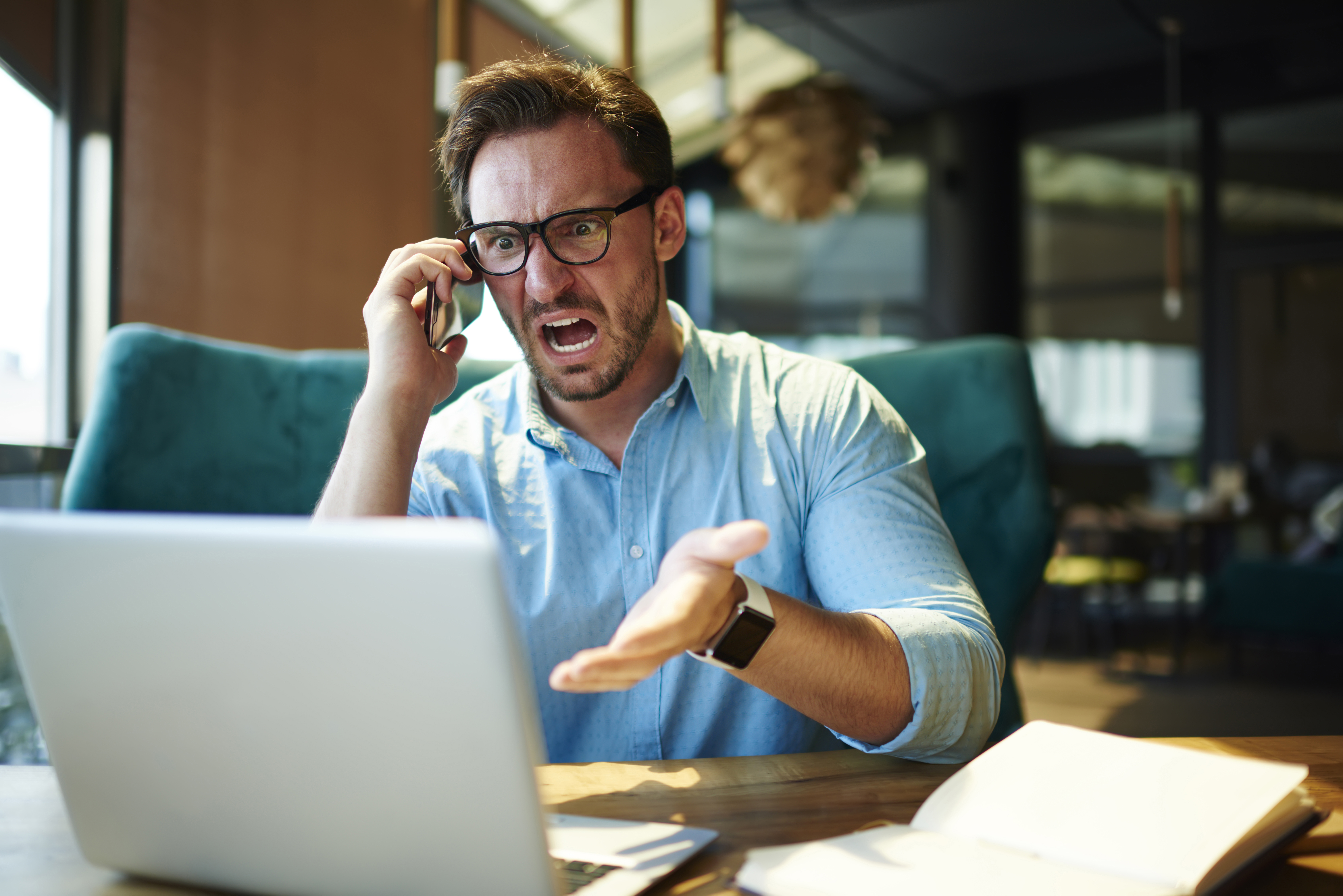 Un homme en colère avec un ordinateur portable devant lui tout en criant au téléphone.│Source : Shutterstock