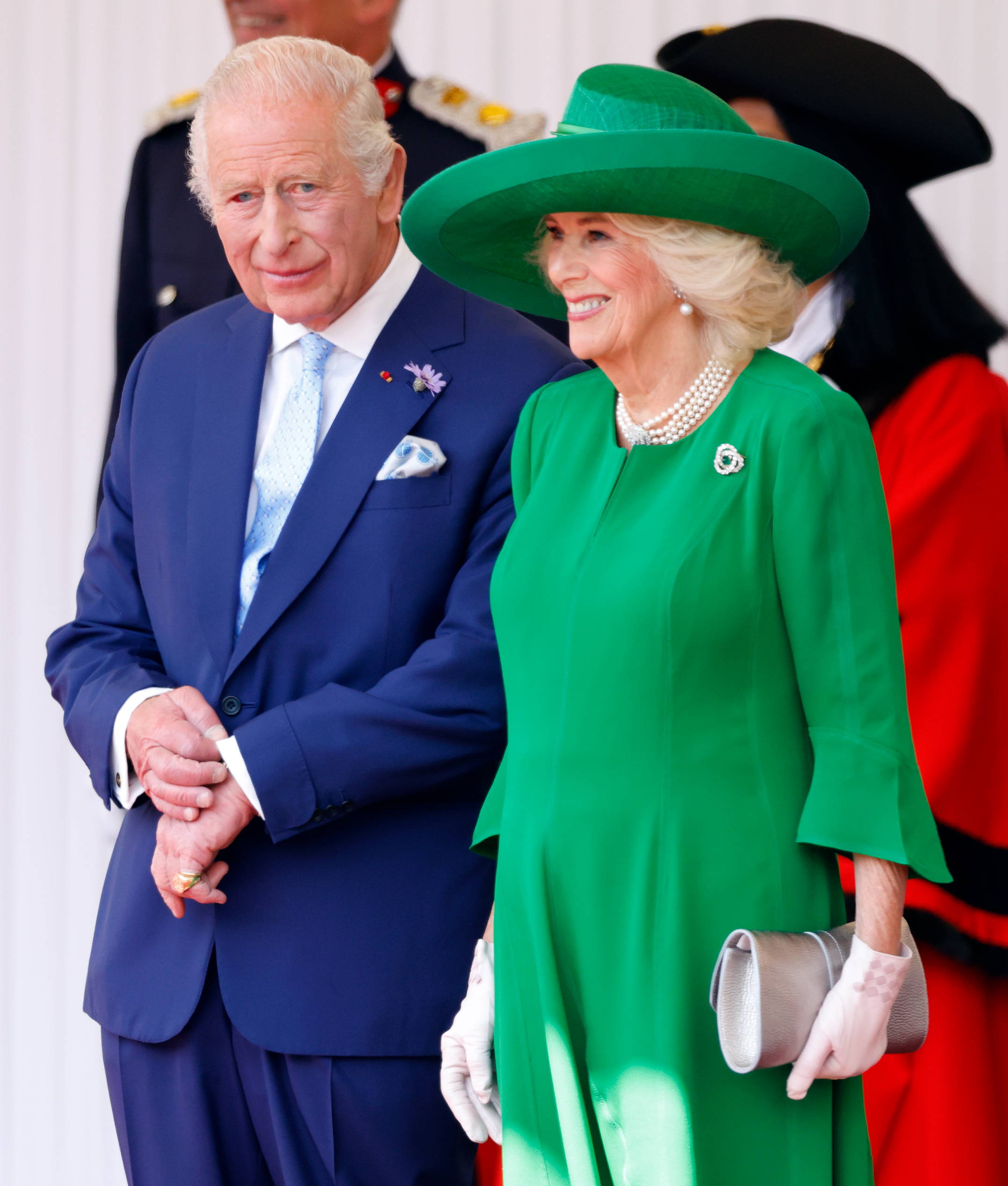 Le roi Charles III et la reine Camilla assistent à la cérémonie officielle d'accueil du président français Emmanuel Macron et de Brigitte Macron à l'occasion de leur visite d'État au Royaume-Uni en 2025. Le roi, vêtu de bleu roi et présentant un œil rouge, se tient aux côtés de son épouse, resplendissante dans une robe vert vif et des perles, sur l'estrade royale à Windsor.