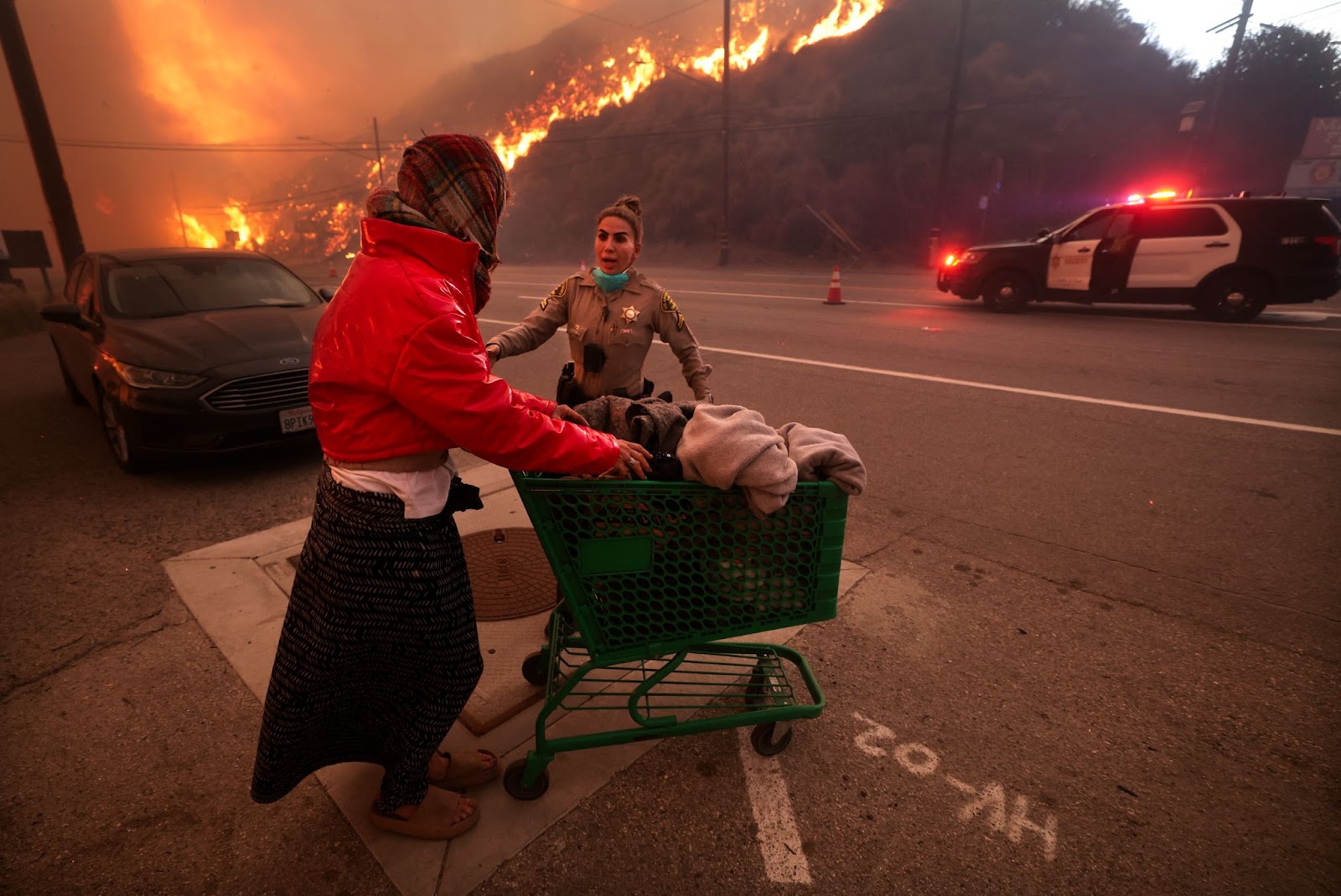 Un officier de police escorte une femme sans-abri loin de Pacific Coast Highway et Topanga Canyon Boulevard alors que l'incendie des Palisades fait rage dans les collines de Los Angeles, Californie, le mardi 7 janvier 2025. | Source : Getty Images