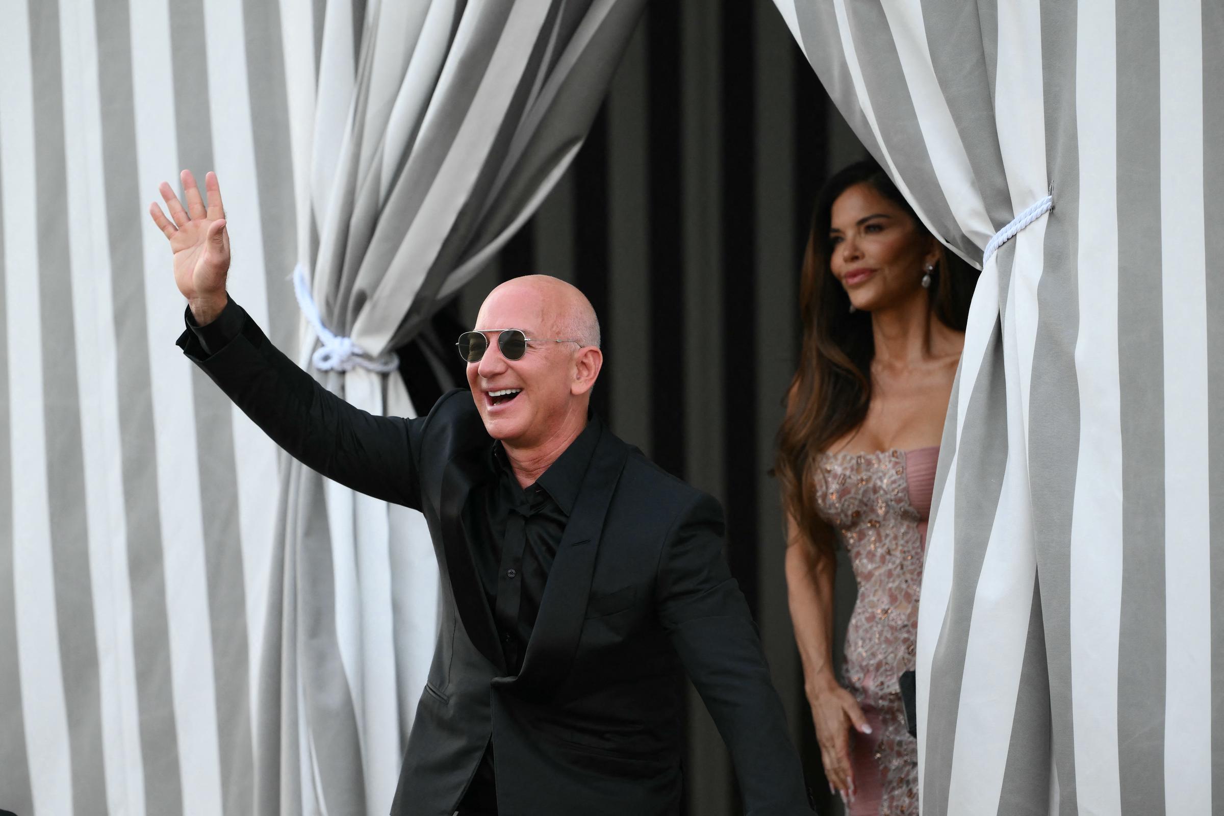 Jeff Bezos rayonne et salue énergiquement les badauds, vêtu d'un costume noir impeccable et de lunettes de soleil aviateur, tandis que Lauren Sánchez se tient gracieusement derrière lui dans une robe chatoyante de couleur blush avec des perles complexes, ses longs cheveux coiffés en vagues lâches tandis qu'elle regarde à l'extérieur, entre d'élégants rideaux rayés.