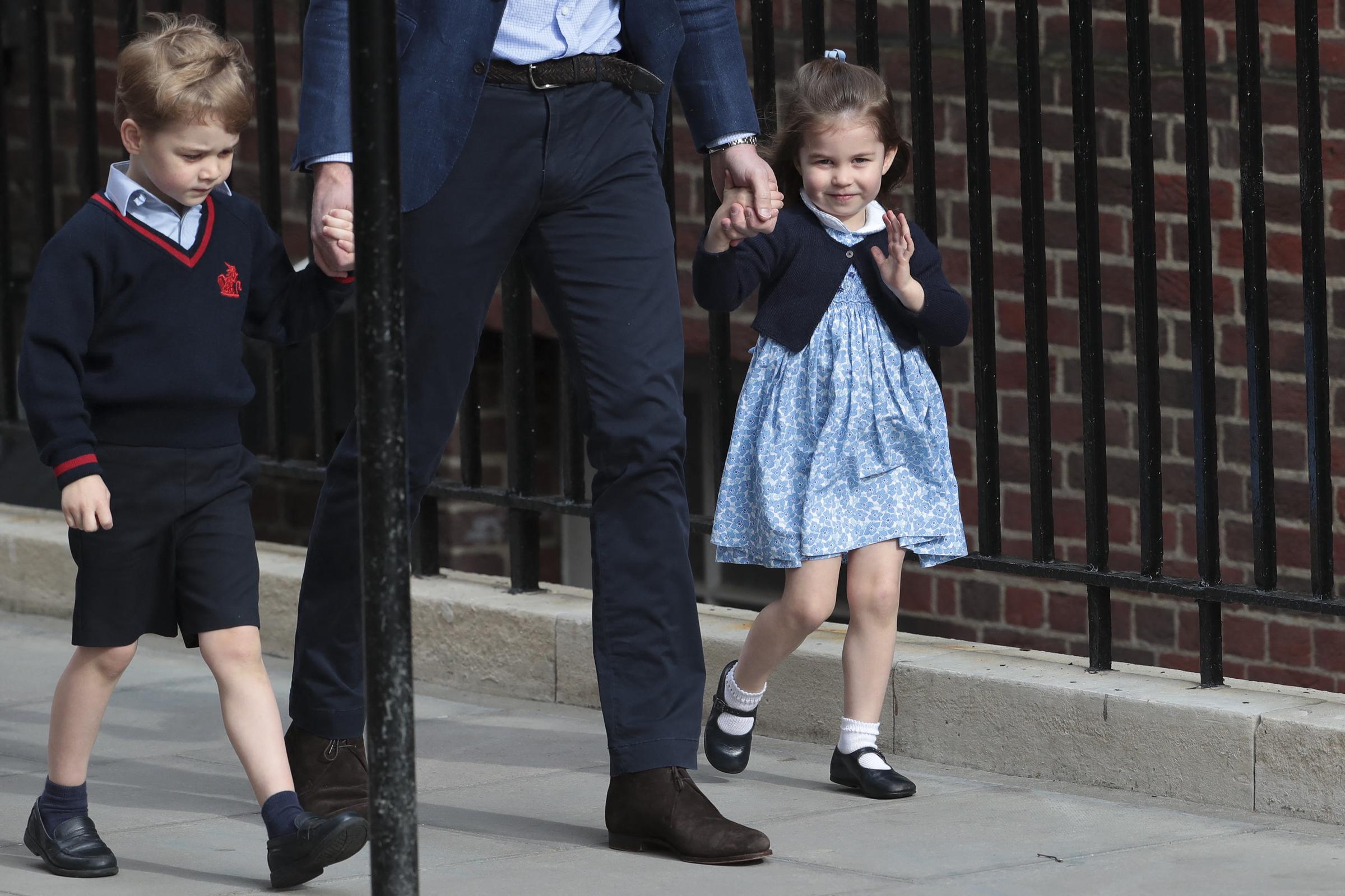 La princesse Charlotte de Cambridge fait un petit signe adorable aux passants devant l'aile Lindo de l'hôpital St Mary's, le 23 avril 2018, à Londres, en Angleterre, alors qu'elle arrive avec son papa, le prince William, et son frère, le prince George, pour rencontrer son petit frère qui vient de naître. La jeune princesse est ravissante dans sa robe-chemise à fleurs bleues et blanches, associée à un cardigan bleu marine, des chaussettes blanches et des chaussures Mary Jane, tandis qu'elle tient la main de son père. | Source : Getty Images