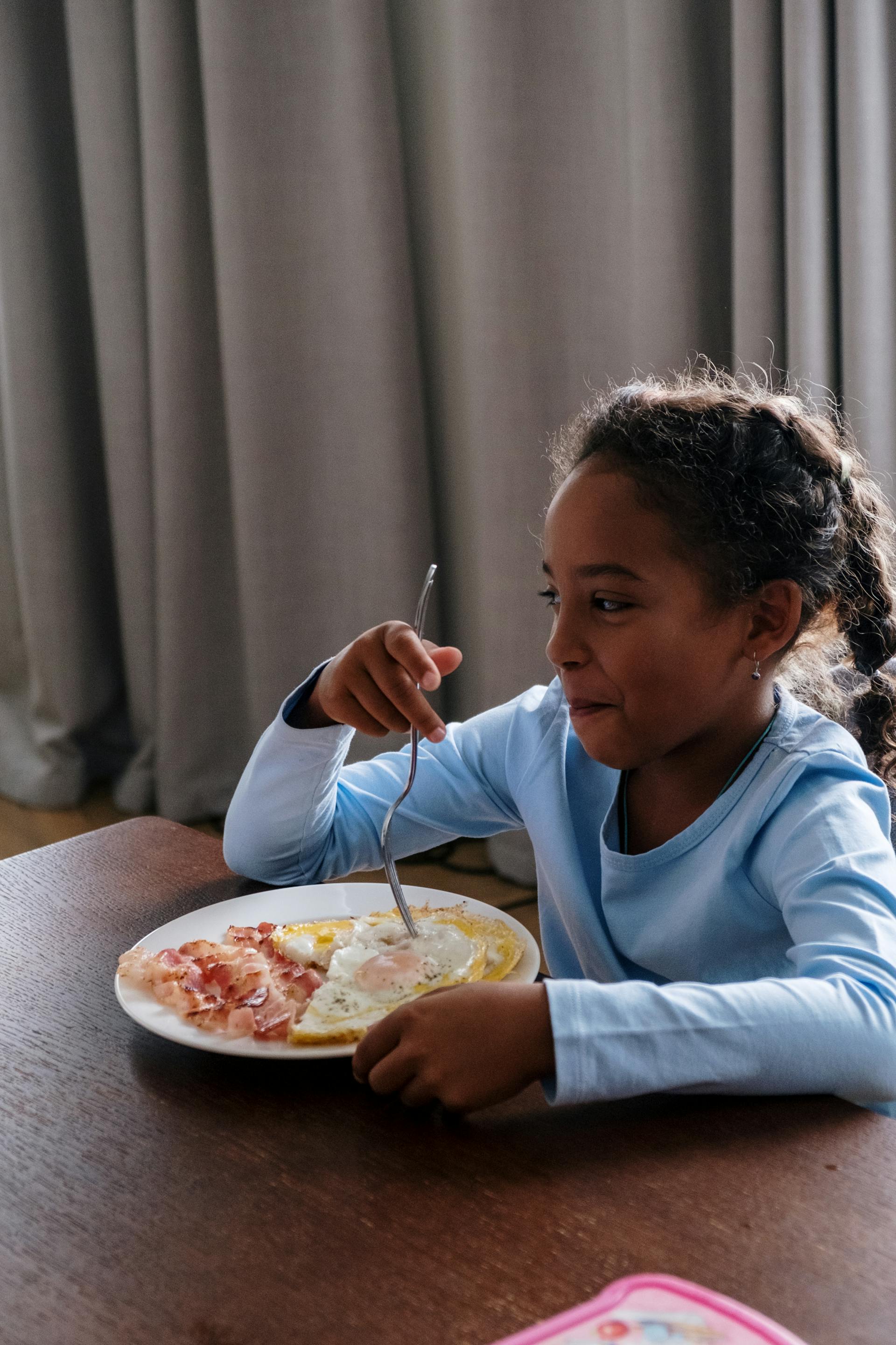 Une petite fille prenant son petit déjeuner essaie de réprimer son rire en regardant quelqu'un | Source : Pexels