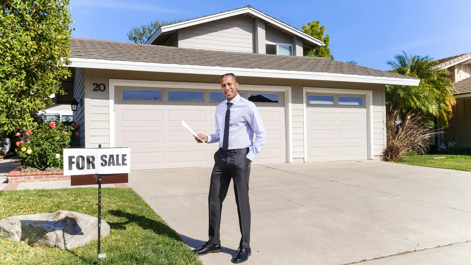 Un homme souriant se tient devant une maison avec un panneau à vendre | Source : Pexels