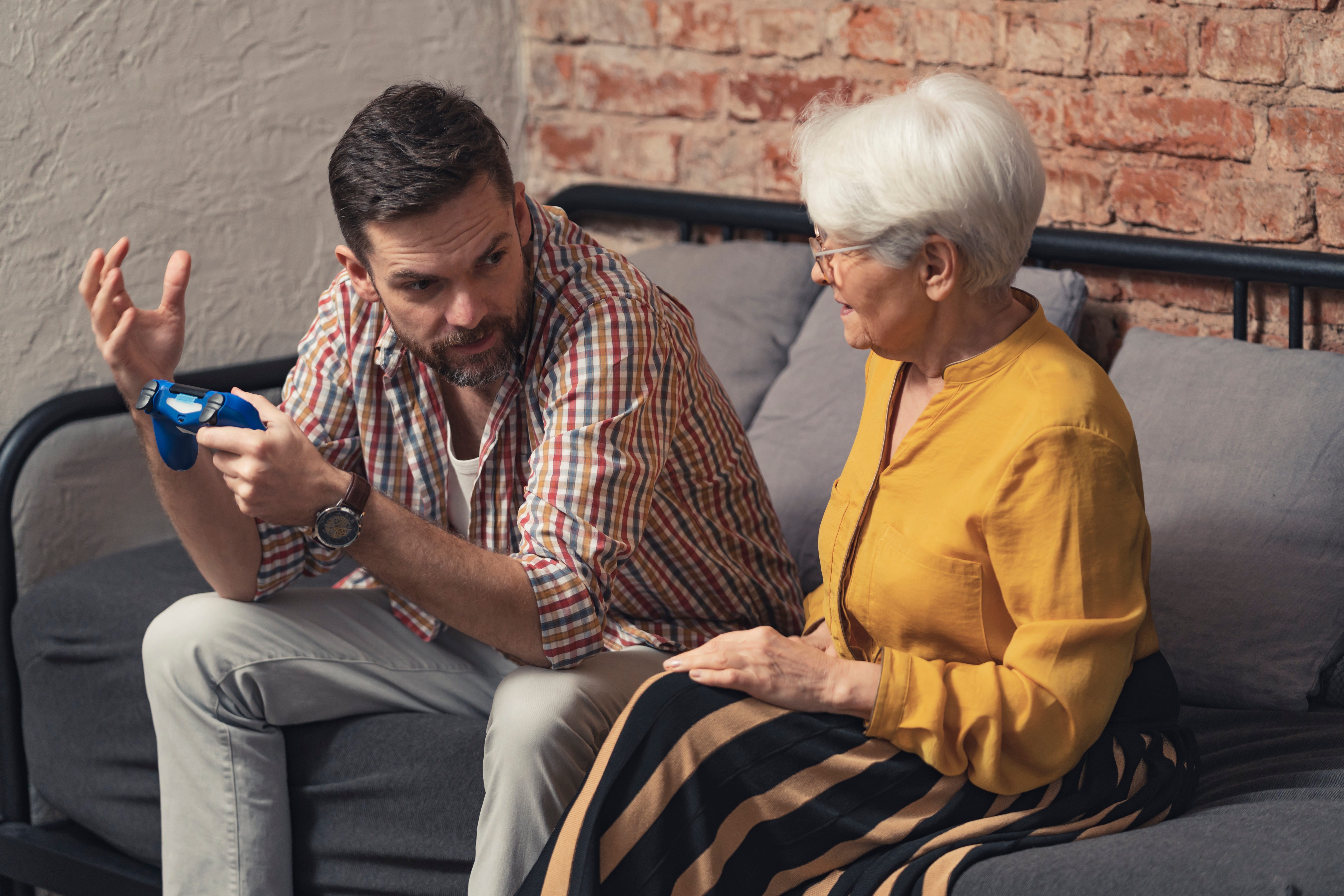 Un homme assis sur un canapé, tenant une manette de jeu vidéo et faisant des gestes avec ses mains en direction d'une dame plus âgée | Source : Shutterstock