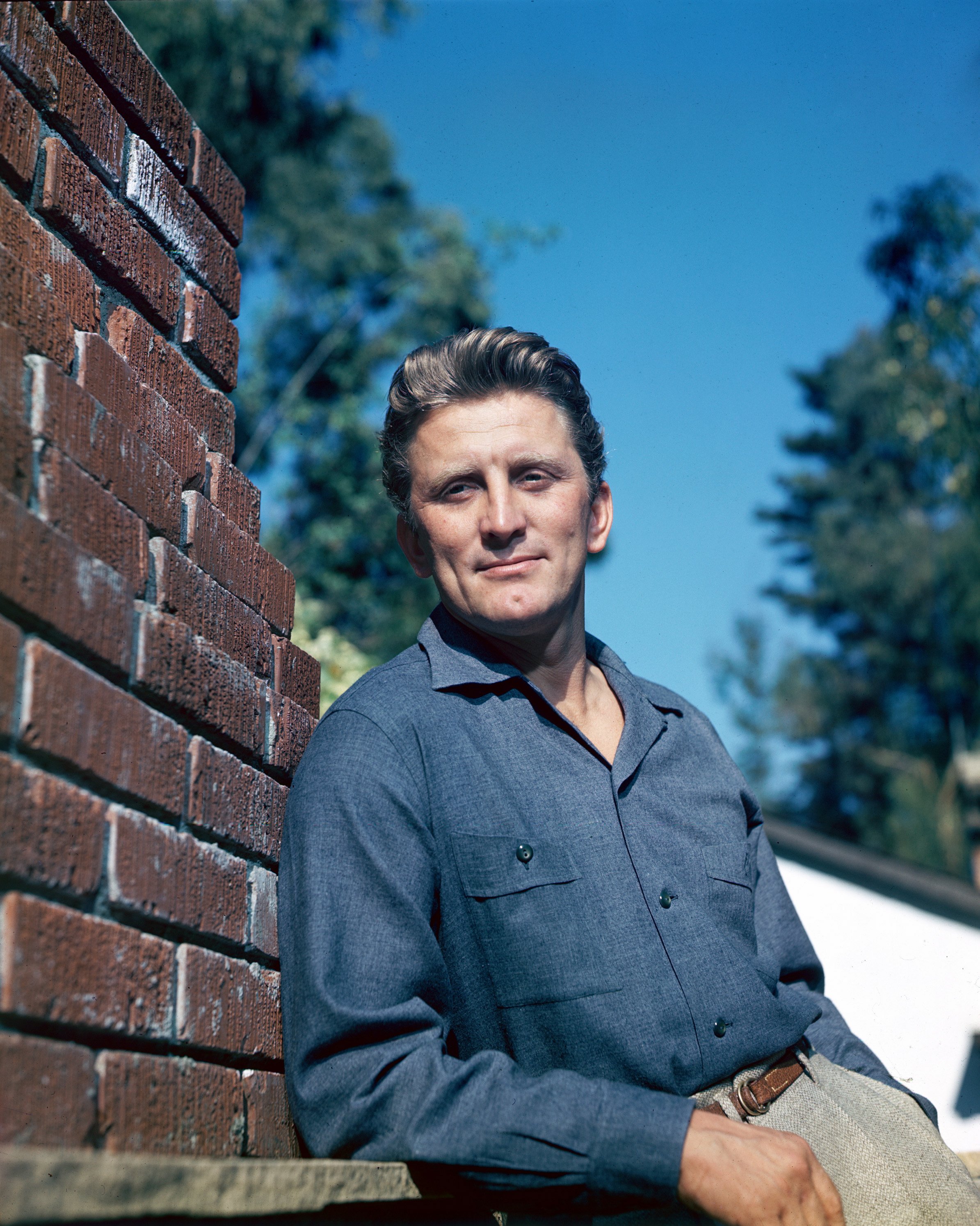 Kirk Douglas photographié adossé au mur portant une chemise bleue associée à un pantalon gris en 1955. / Source : Getty Images