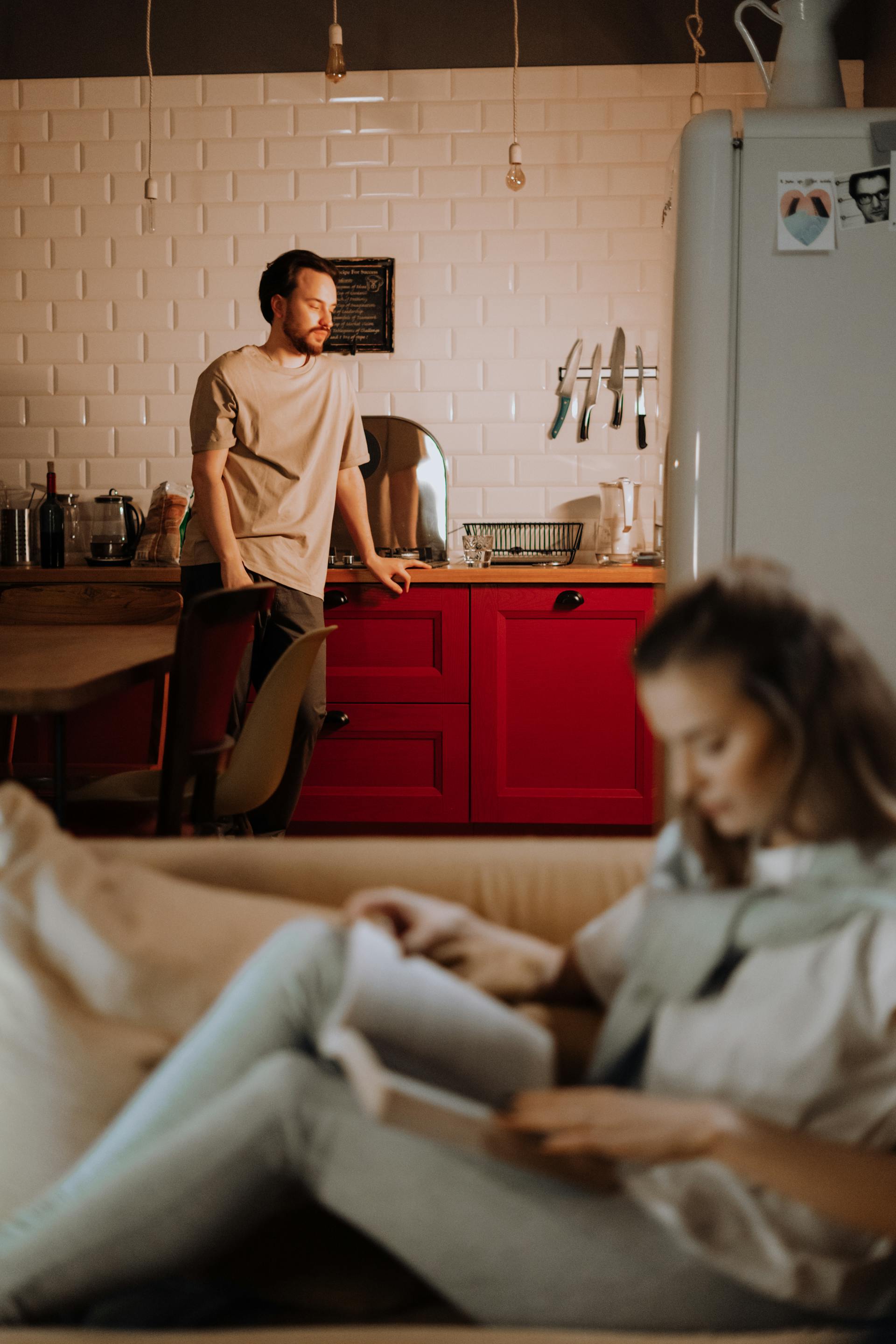 Une femme lit un livre sur le canapé tandis qu'un homme en détresse se tient dans la cuisine | Source : Pexels