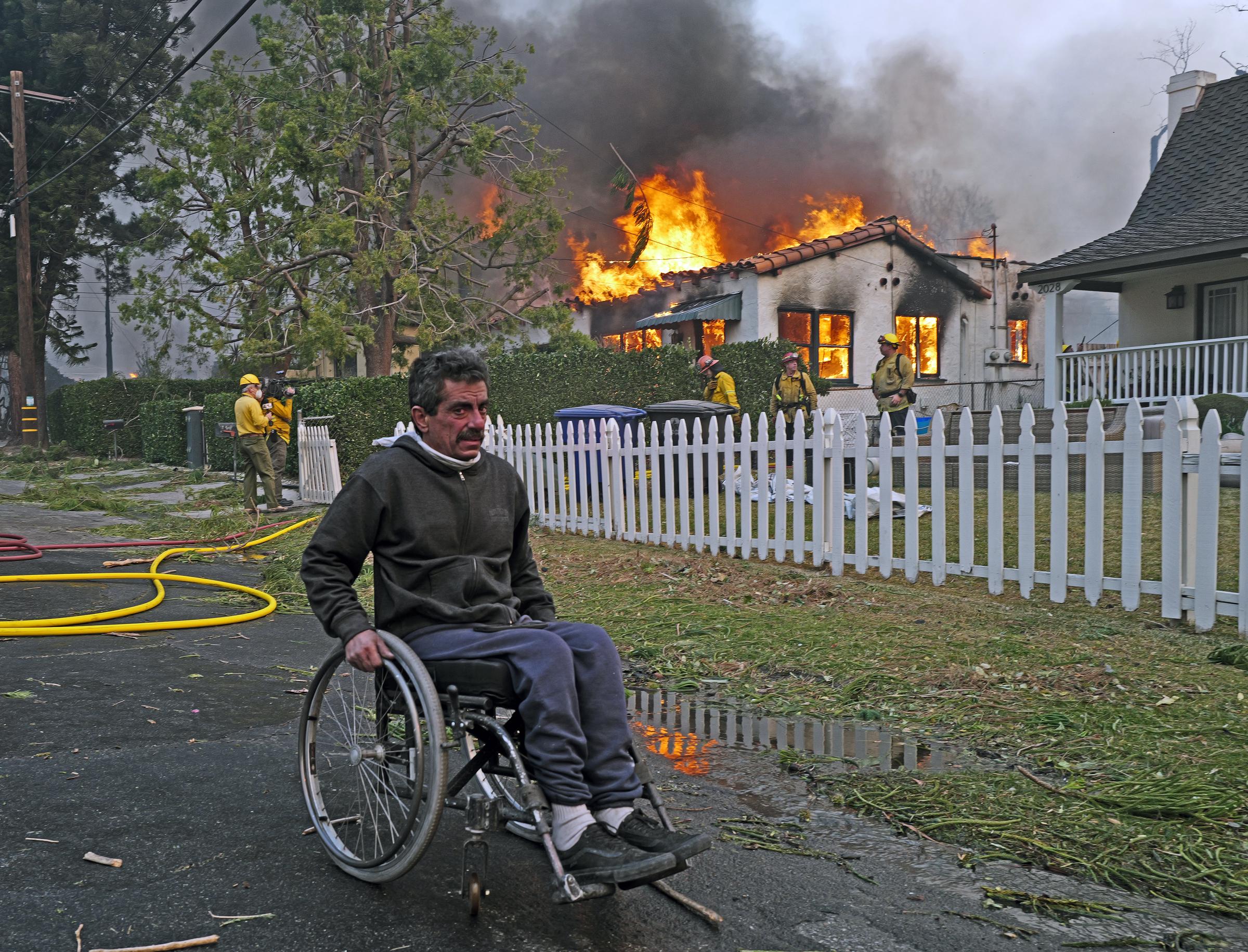 Un homme en fauteuil roulant passe devant une maison en feu suite à l'incendie d'Eaton dans le quartier d'Altadena, le 08 janvier 2025, à Pasadena, en Californie | Source : Getty Images