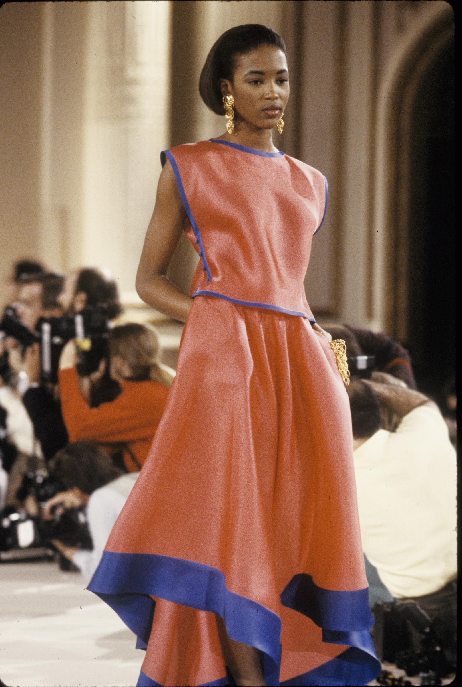 La mannequin Naomi Campbell porte une robe rouge sans manches à jupe ample, rehaussée de bordures bleues, lors du défilé prêt-à-porter printemps-été 1989 d'Oscar de la Renta à New York I Source : Getty Images