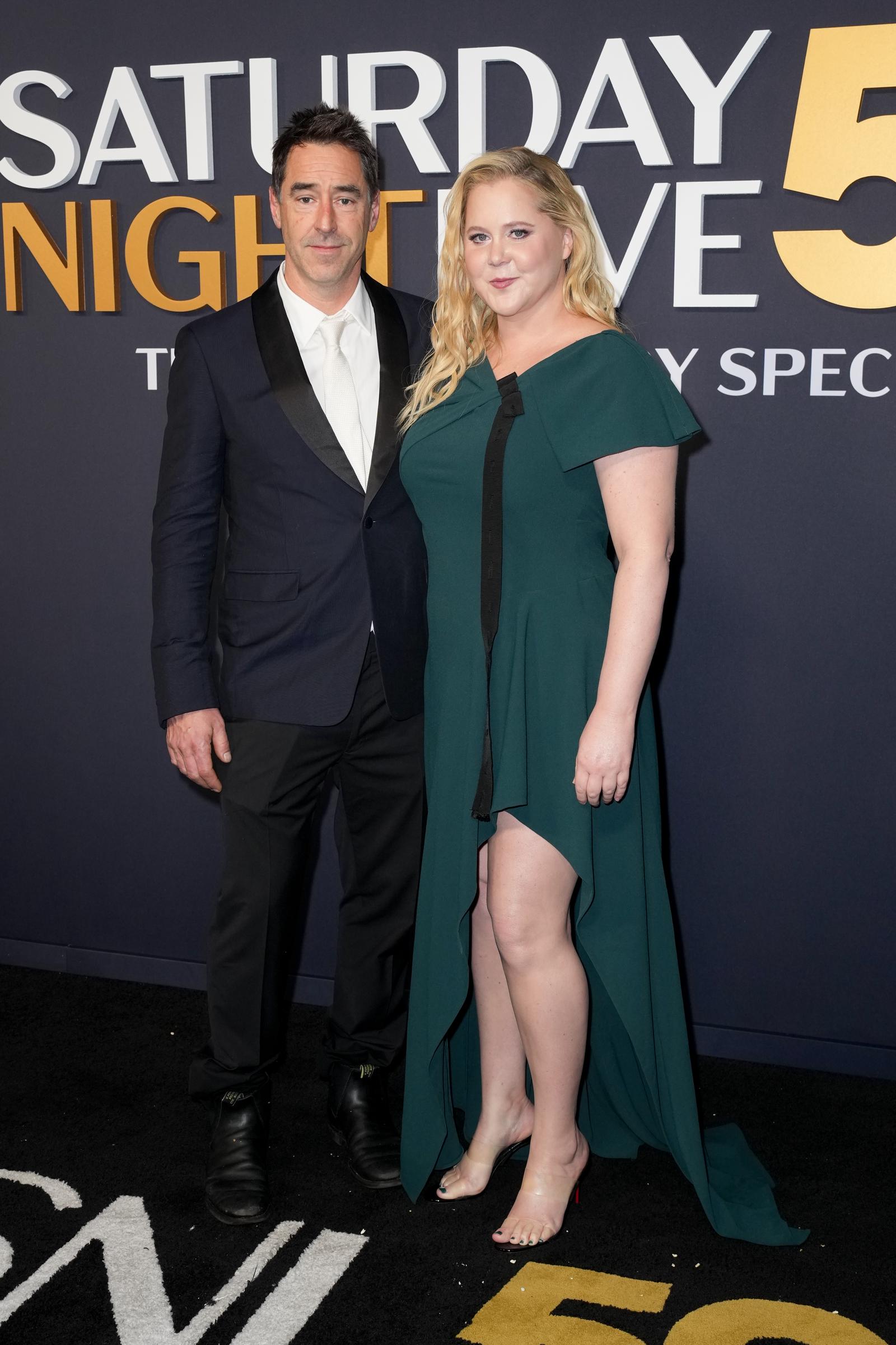 Lors du SNL 50, le 16 février 2025, Amy Schumer portait une robe verte à manches cape et fente haute, posant aux côtés de son mari Chris Fischer, qui portait un smoking noir classique avec une cravate blanche.