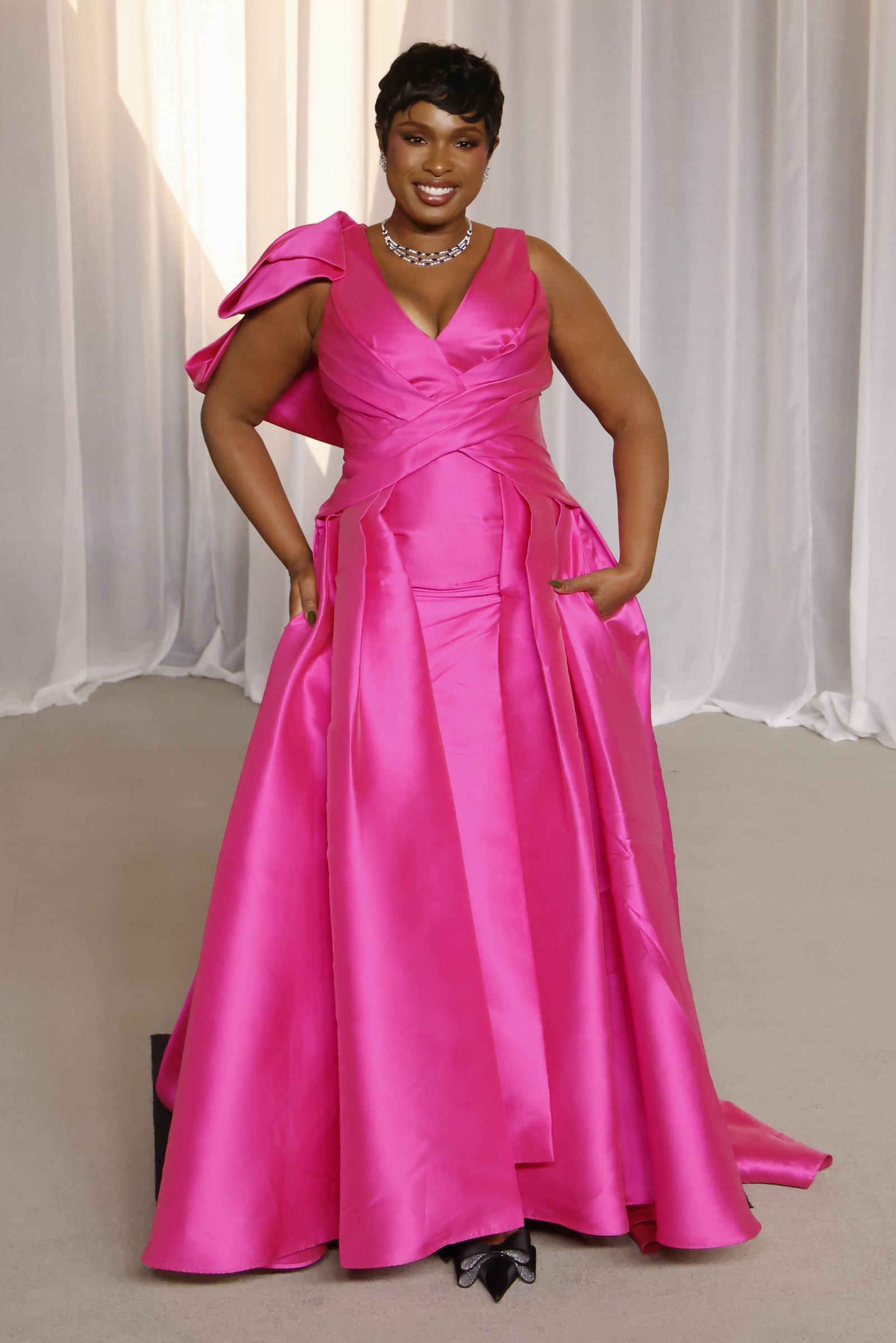 Jennifer Hudson attire tous les regards dans une robe en satin rose vif éclatante, avec un décolleté en V plongeant et des drapés sculptés le long du corsage. Un nœud asymétrique et volumineux vient rehausser une épaule, tandis que la jupe ample, qui effleure le sol, est dotée de poches intégrées pour un charme tout en simplicité. La lauréate d’un EGOT complète ce look époustouflant avec un collier ras-du-cou en diamants étincelants, des escarpins à bout pointu ornés de nœuds en cristal et une coupe de cheveux pixie ultra-épurée.