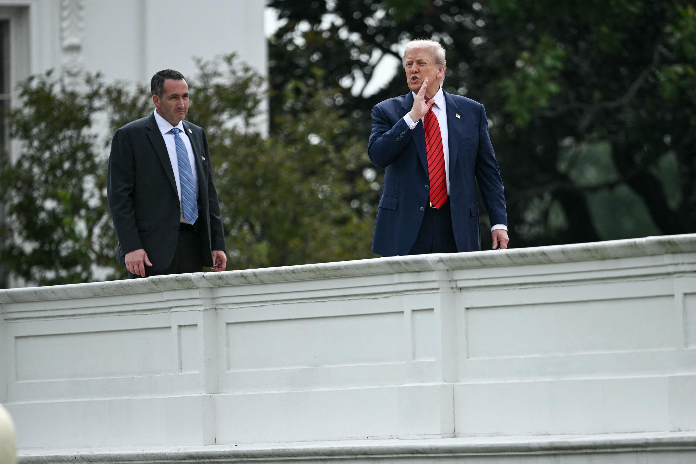 Donald Trump se tient sur le toit de la Maison Blanche, une main levée vers sa bouche comme s'il appelait ou s'adressait à quelqu'un en bas, avec un membre de la sécurité à ses côtés.