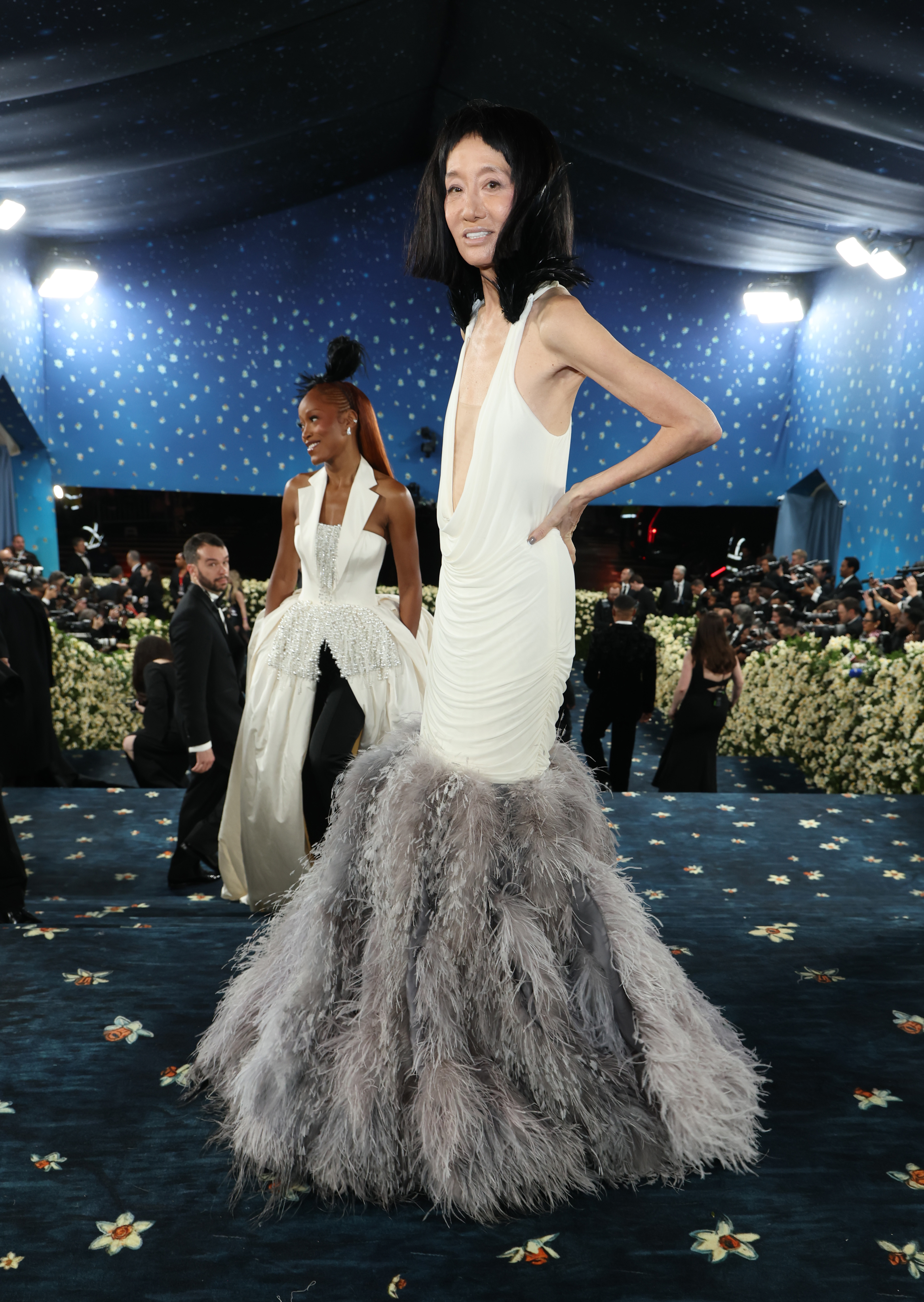 Entourée d'une mer de caméras et de décorations florales, Vera Wang pose à mi-pas dans sa robe texturée tandis que Keke Palmer la suit, toutes deux vêtues de looks blancs frappants avec des plumes.