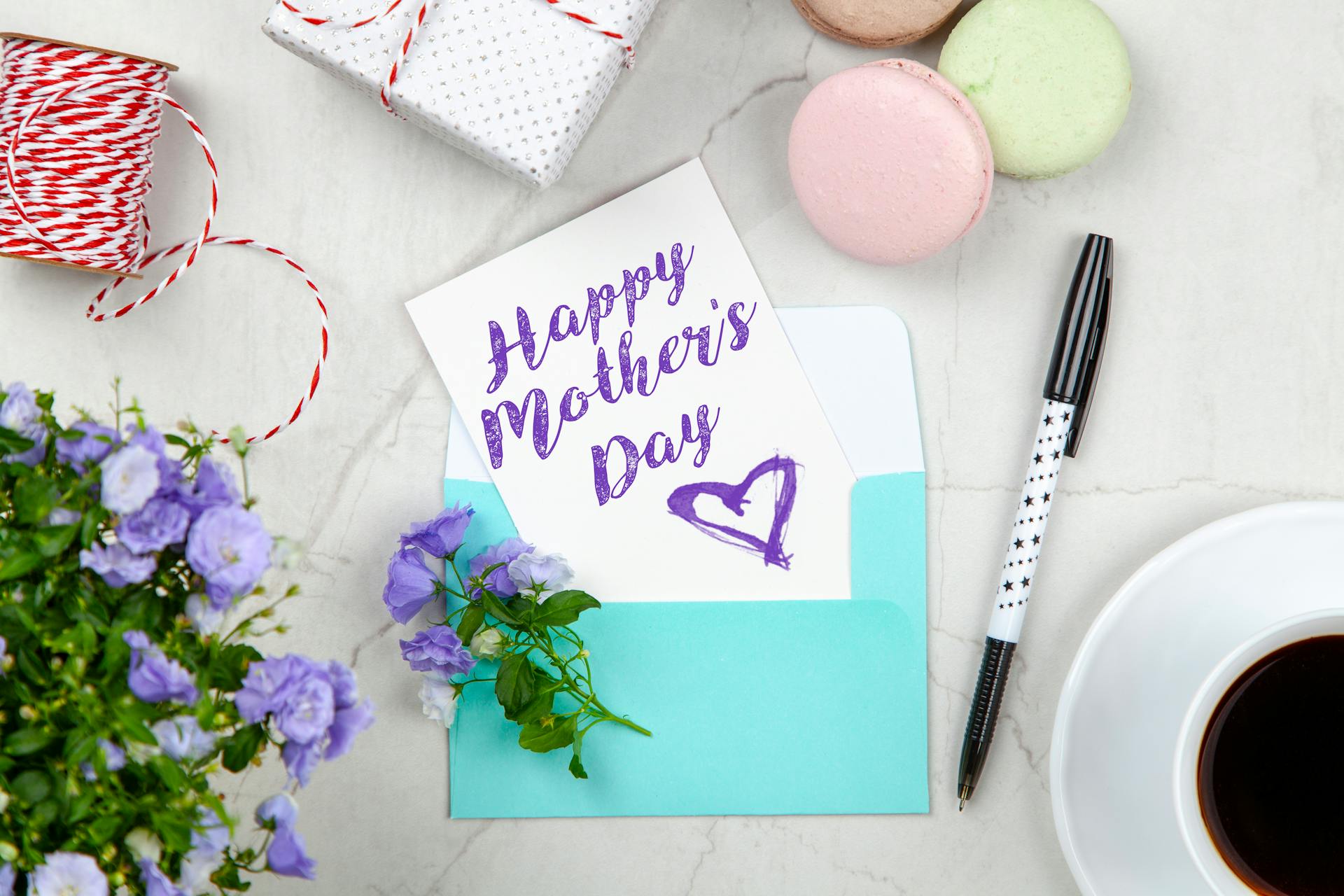 Une carte de fête des mères à côté d'un stylo, de macarons, de fleurs et d'une boîte près d'une tasse à café avec soucoupe | Source : Pexels