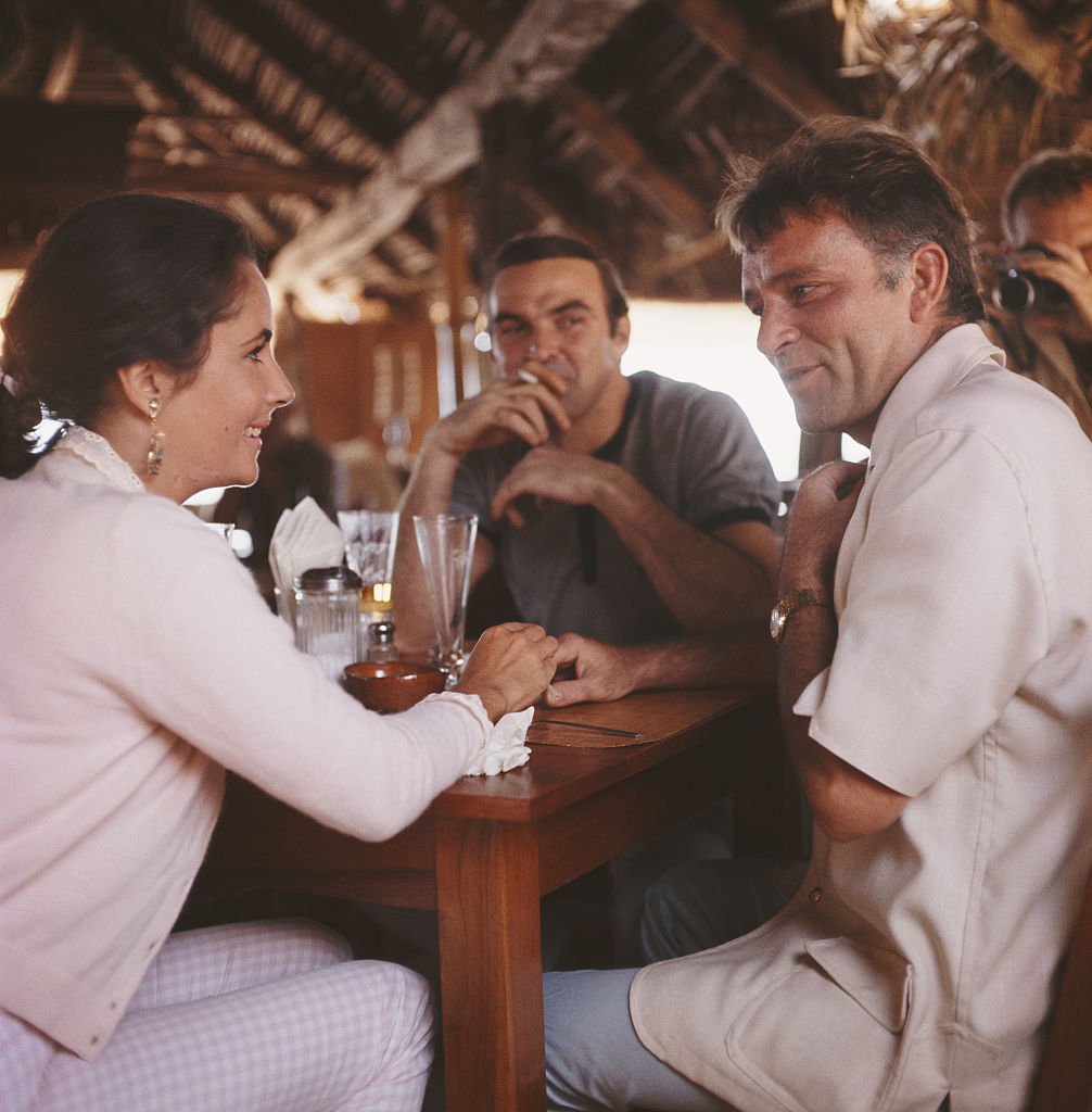 De gauche à droite : les acteurs Elizabeth Taylor (1932 - 2011), Stanley Baker (1928 - 1976) et Richard Burton (1925 - 1984) dans un bar, Puerto Vallarta, Mexique, 12 janvier 1964. Burton est au Mexique pour le tournage du film "La nuit de l'Iguane" de John Huston. | Source : Getty Images