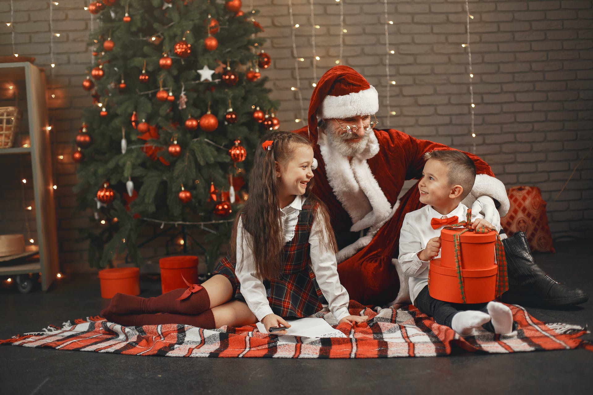 Homme en costume de Père Noël assis avec un garçon et une fille à côté d'un sapin de Noël | Source : Pexels