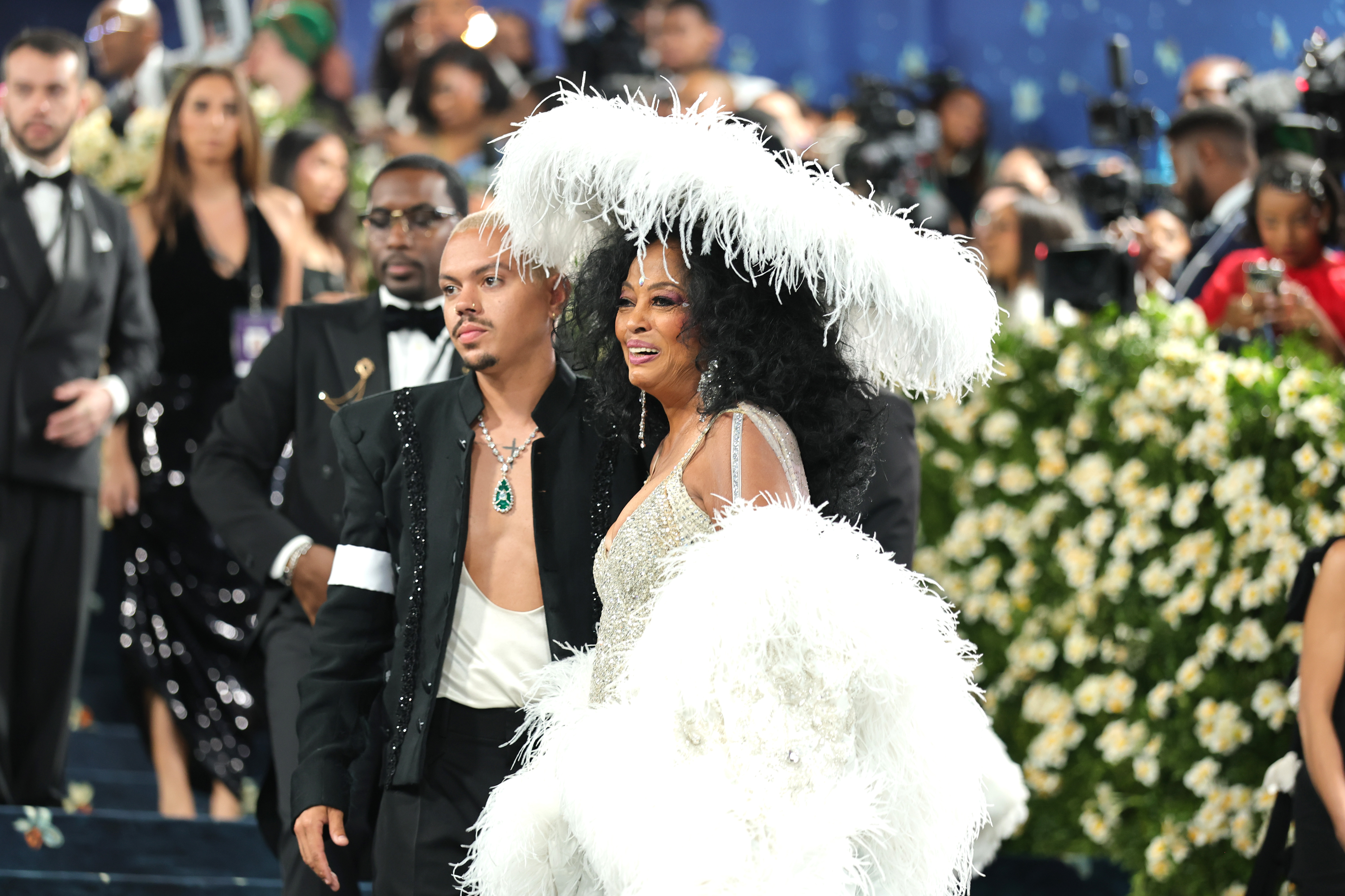 Sur un plan plus rapproché, le décolleté ouvert d'Evan Ross révèle un tatouage en forme de croix sur sa poitrine et un collier avec une goutte d'émeraude frappante reposant contre son cou. À côté de lui, sa mère sourit gentiment, l'expression chaleureuse et fière.