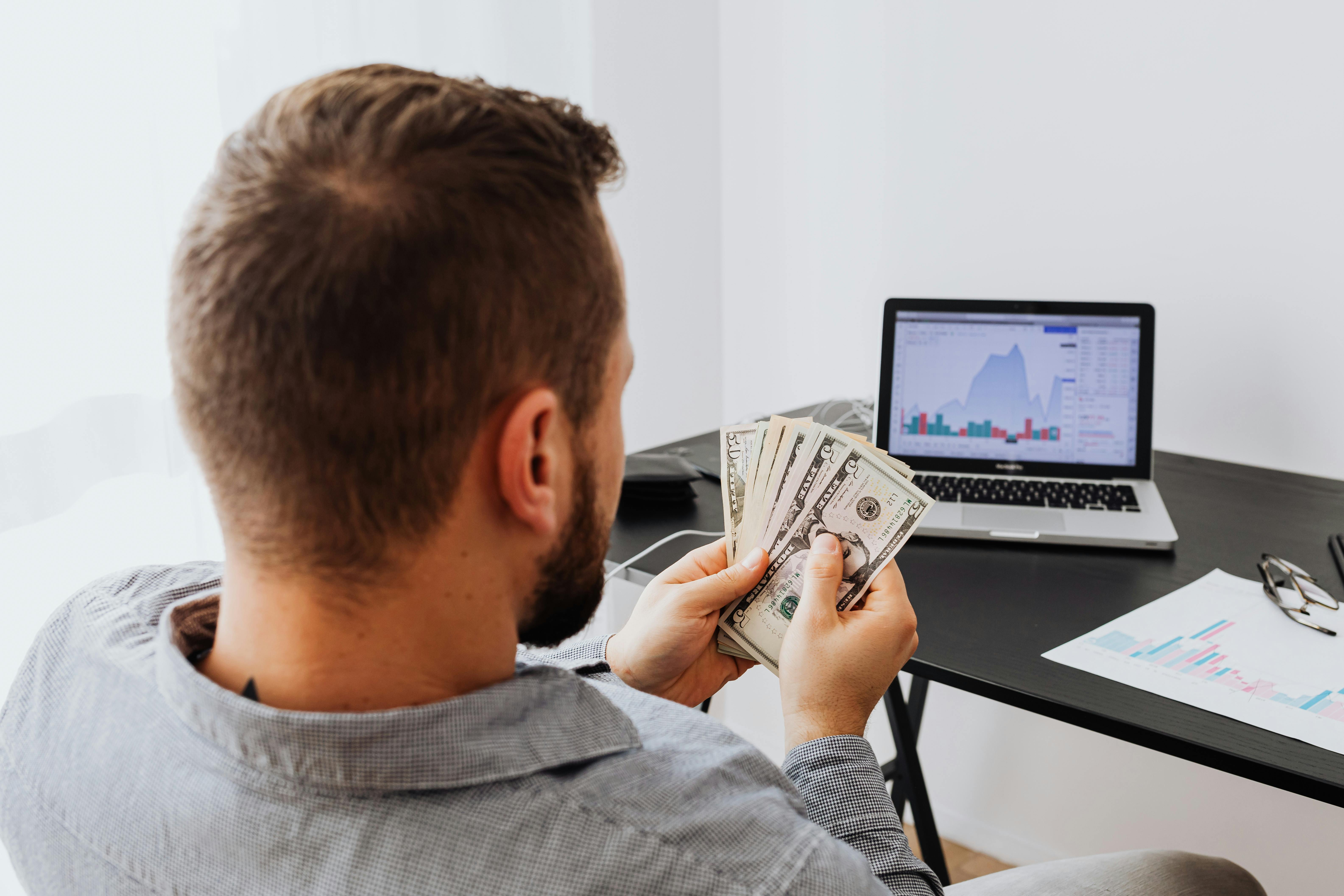 Un homme assis devant un ordinateur portable en train de compter de l'argent | Source : Pexels